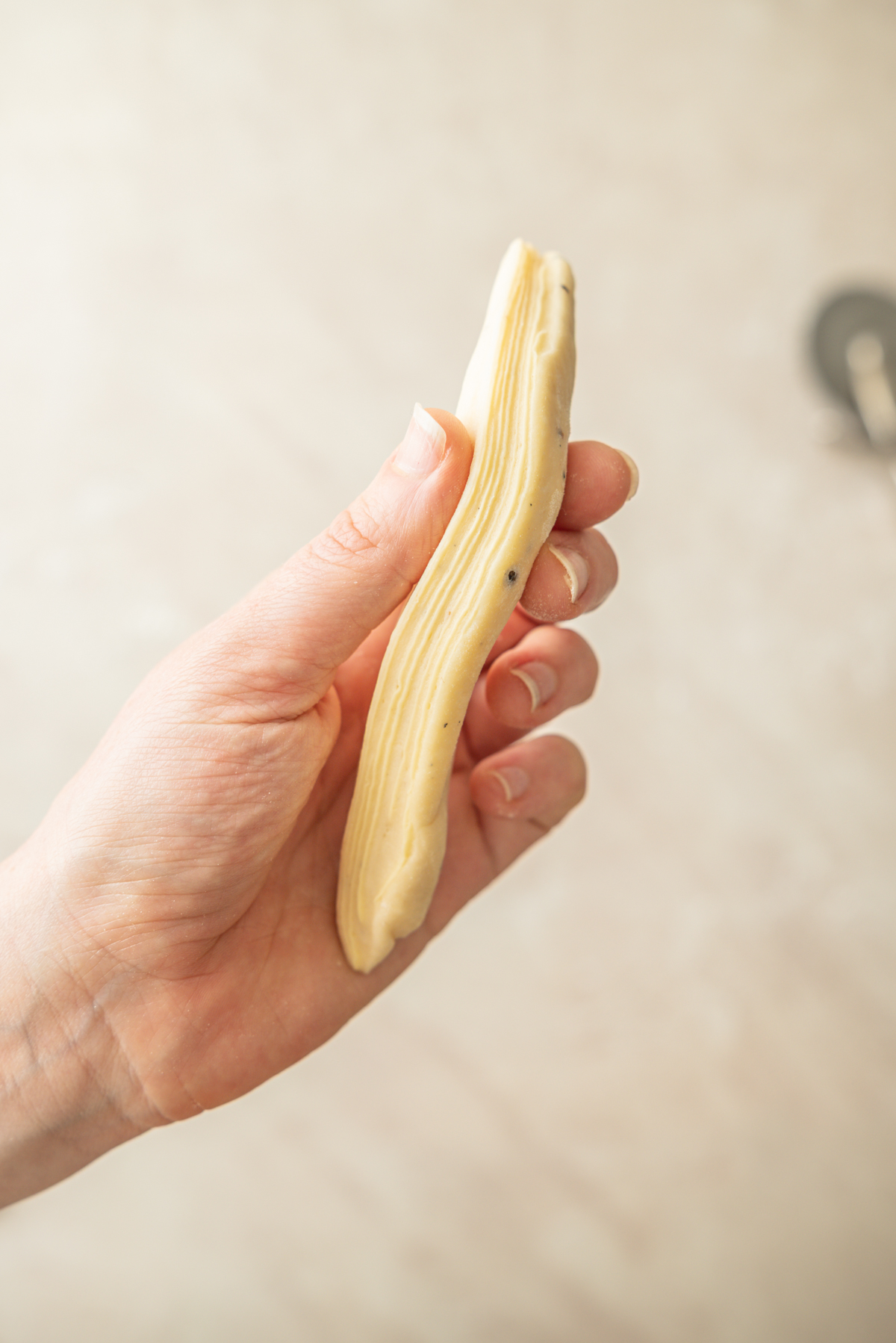 A hand holding a piece of layered pastry dough with visible thin layers, set against a neutral, blurred background.