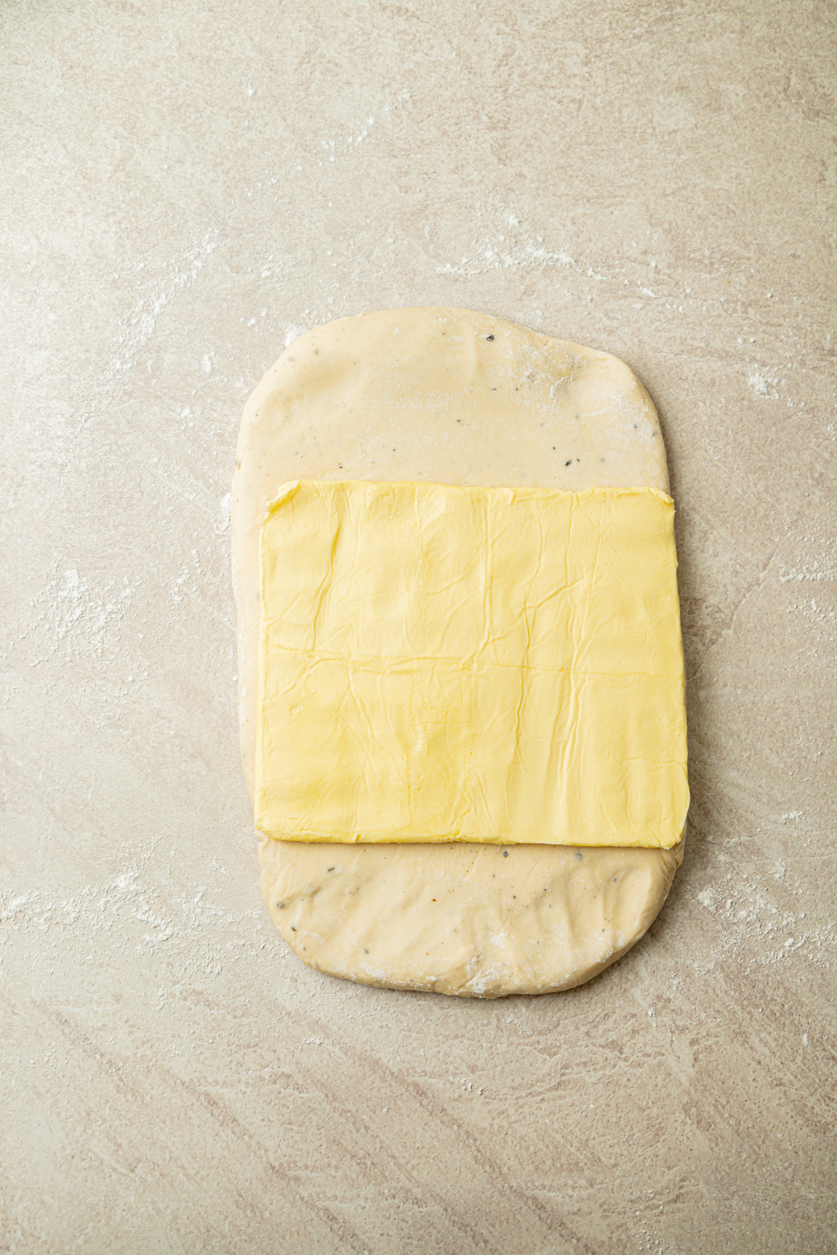 A rectangular slab of butter placed on a rolled-out piece of dough on a floured surface, ready for folding.