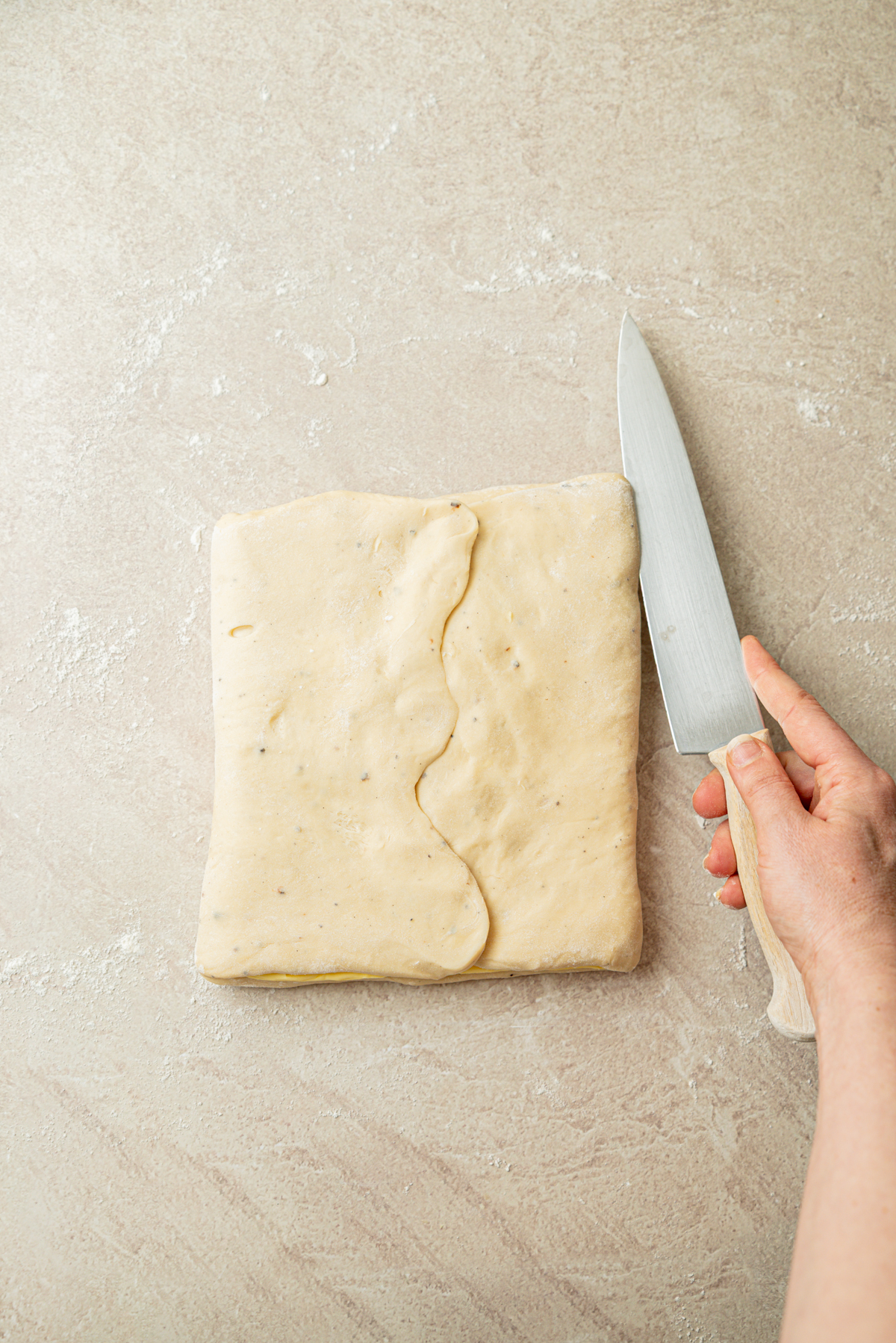 A hand holding a knife next to folded dough on a lightly floured surface, ready to be cut.