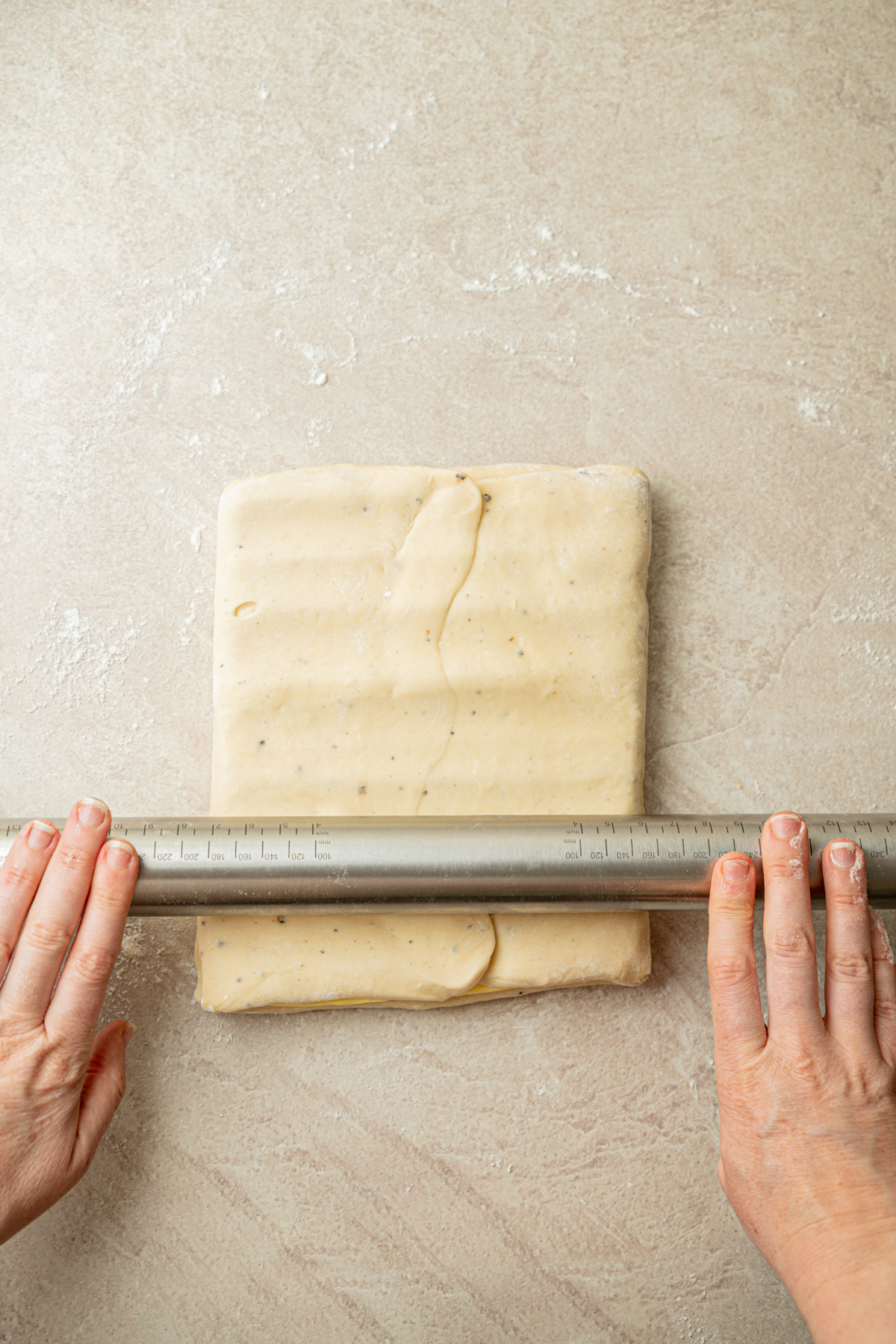 A person uses a metal ruler to measure a rectangular slab of dough on a floured surface.