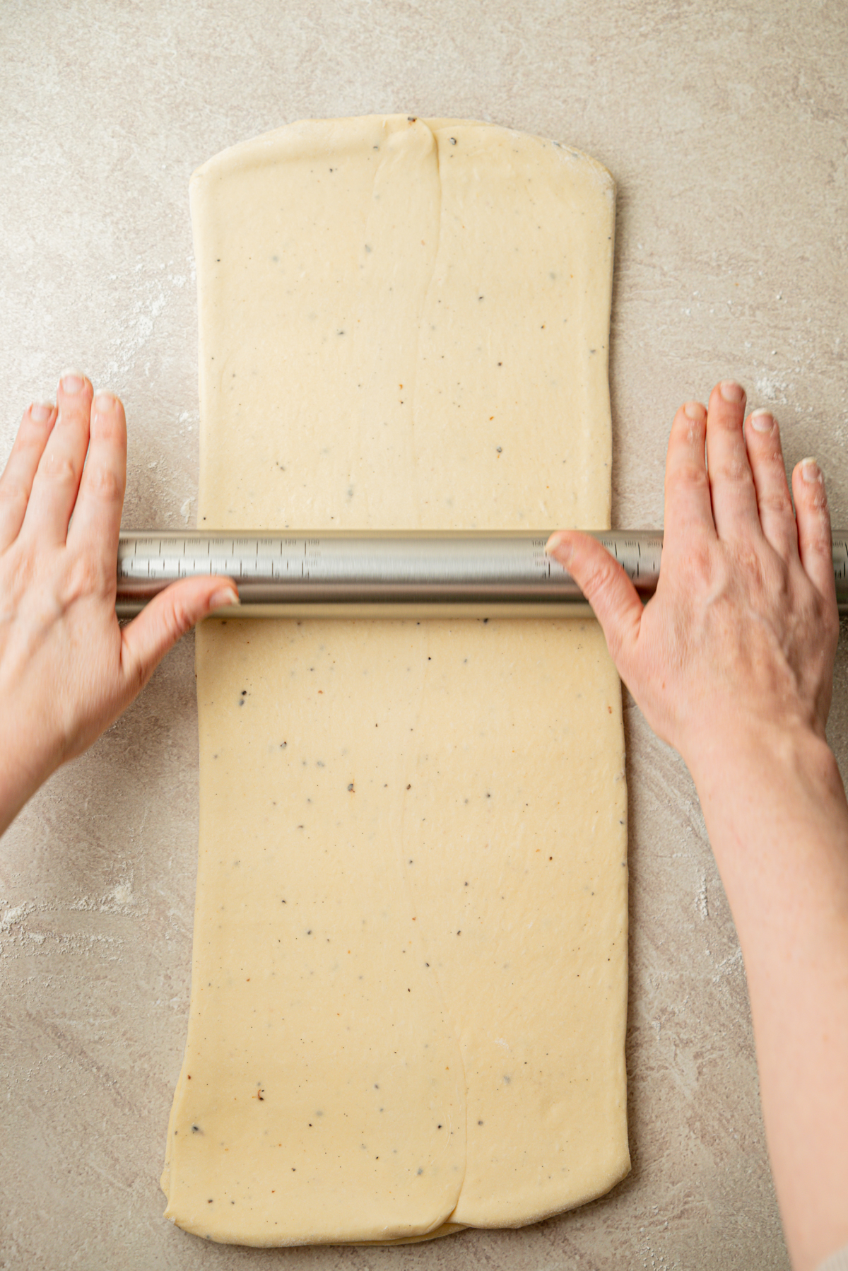 Hands rolling out rectangular dough with a metal rolling pin on a light countertop.