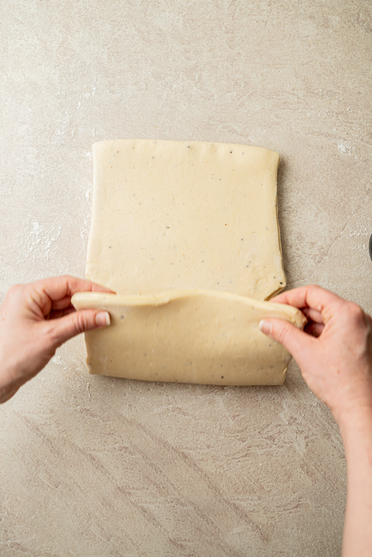 Two hands fold a rectangular sheet of dough on a lightly floured, beige surface, preparing it for baking.