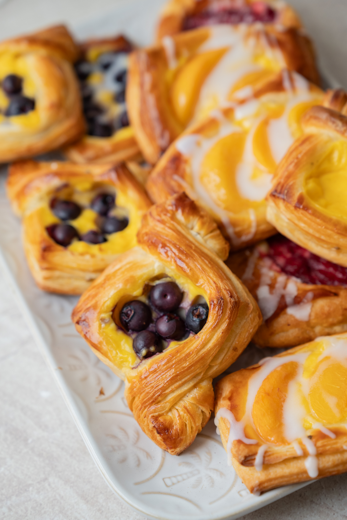 A plate of assorted fruit danishes topped with blueberries and peach slices, drizzled with white icing, displayed on a white tray.