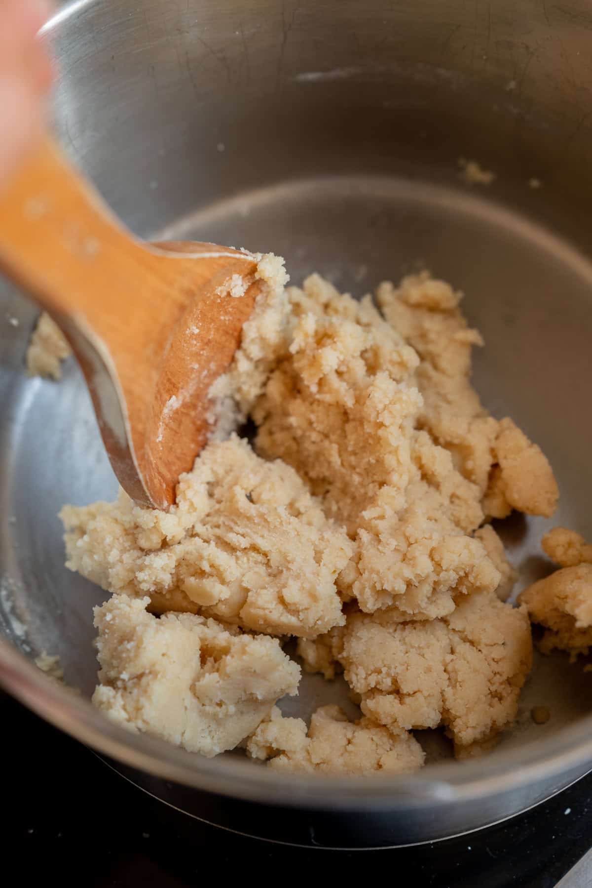 A wooden spoon stirs thick, crumbly homemade marzipan dough in a metal mixing bowl.