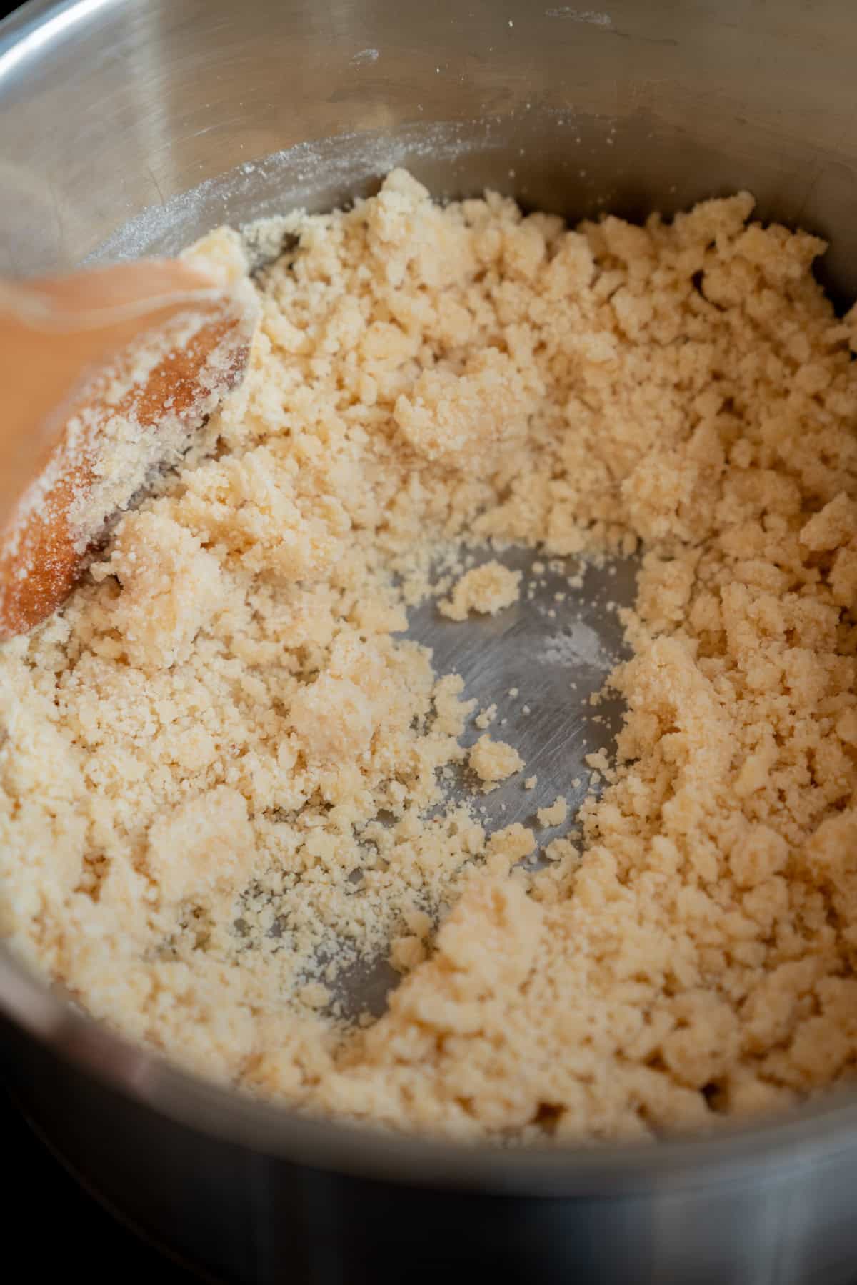 A close-up of a saucepan with crumbly Homemade Marzipan dough being stirred by a wooden spoon, showing a partially mixed texture.