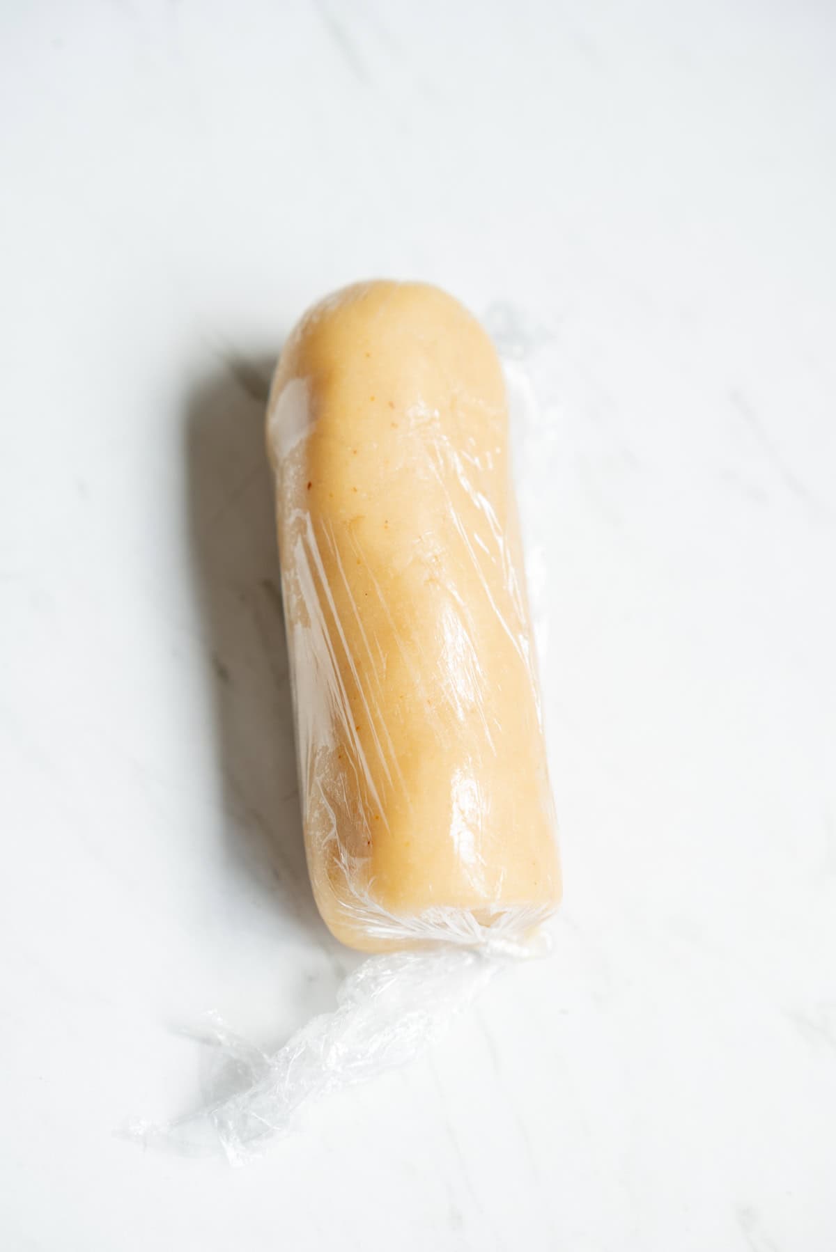 A roll of light beige homemade marzipan dough wrapped tightly in plastic wrap, placed on a white marble surface.
