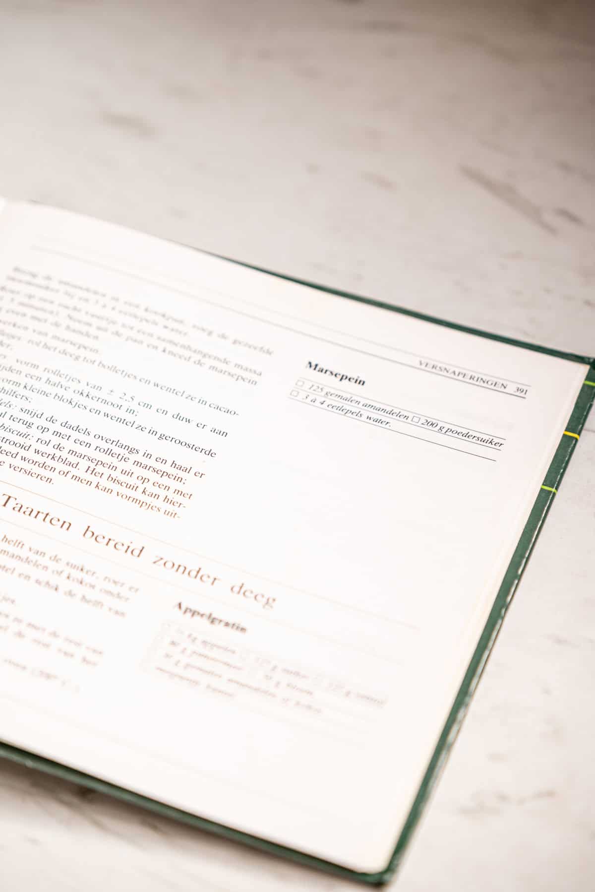 An open cookbook on a marble surface shows a homemade marzipan recipe in Dutch, highlighting ingredients and instructions in black text on white pages with green and red accents.
