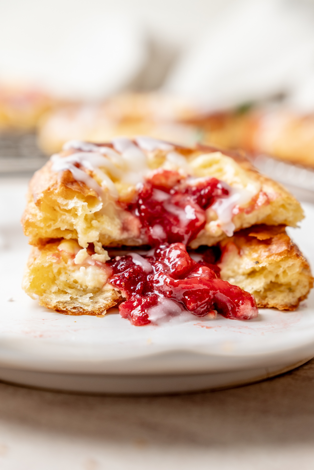 A close-up of a pastry split open on a plate, revealing a bright red cherry filling and drizzled with white icing. The background is softly blurred.