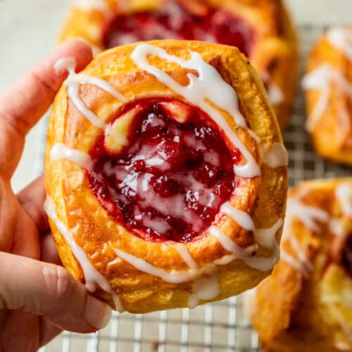 A hand holds a raspberry danish pastry with white icing drizzle. More pastries are visible on a cooling rack in the background.