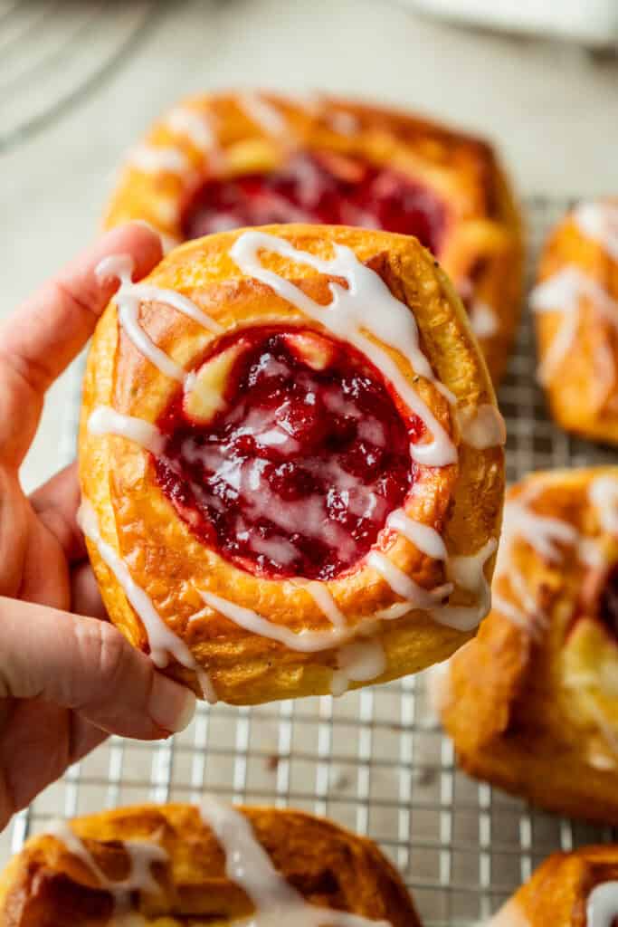 A hand holds a raspberry danish pastry with white icing drizzle. More pastries are visible on a cooling rack in the background.