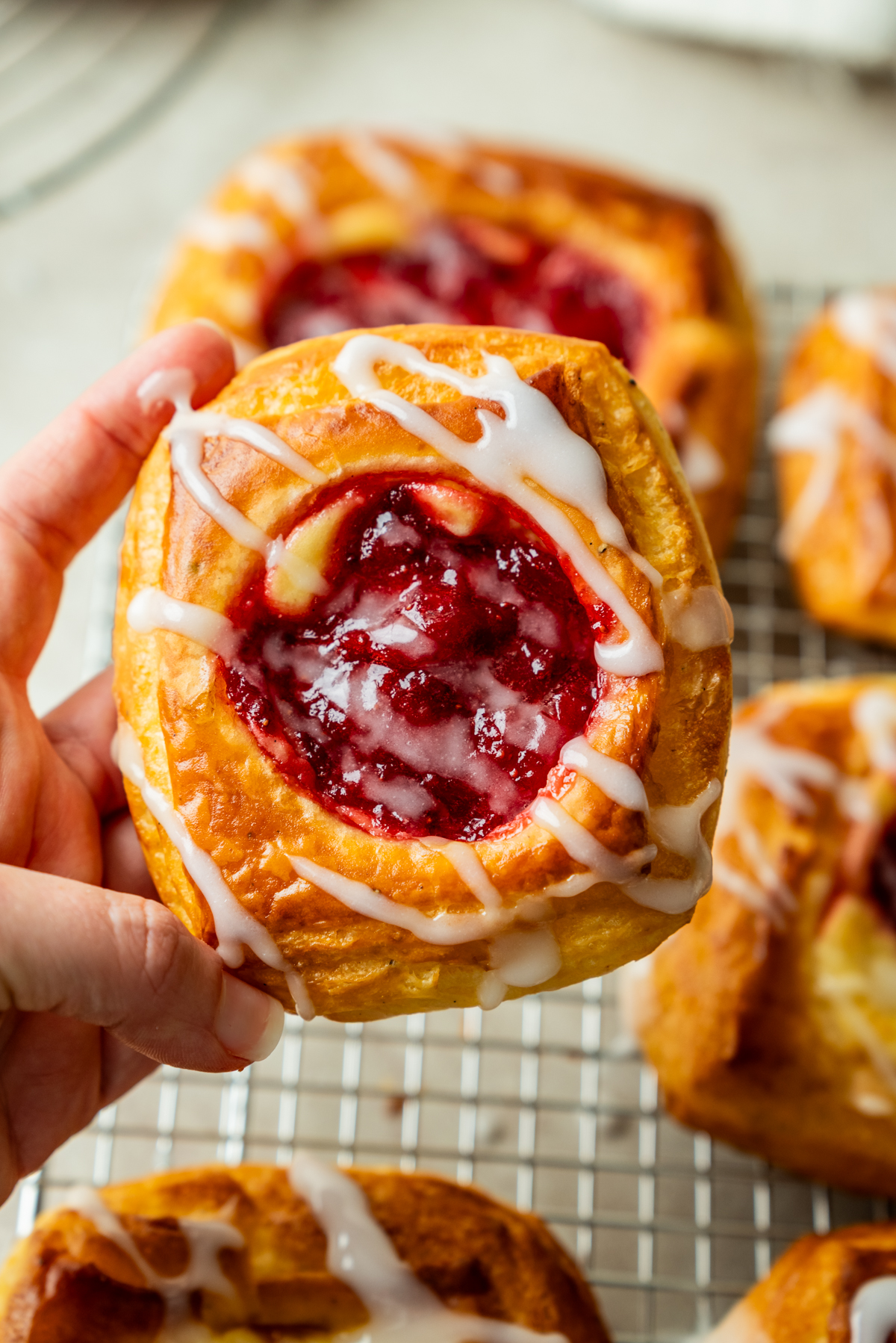 A hand holds a raspberry danish pastry with white icing drizzle. More pastries are visible on a cooling rack in the background.