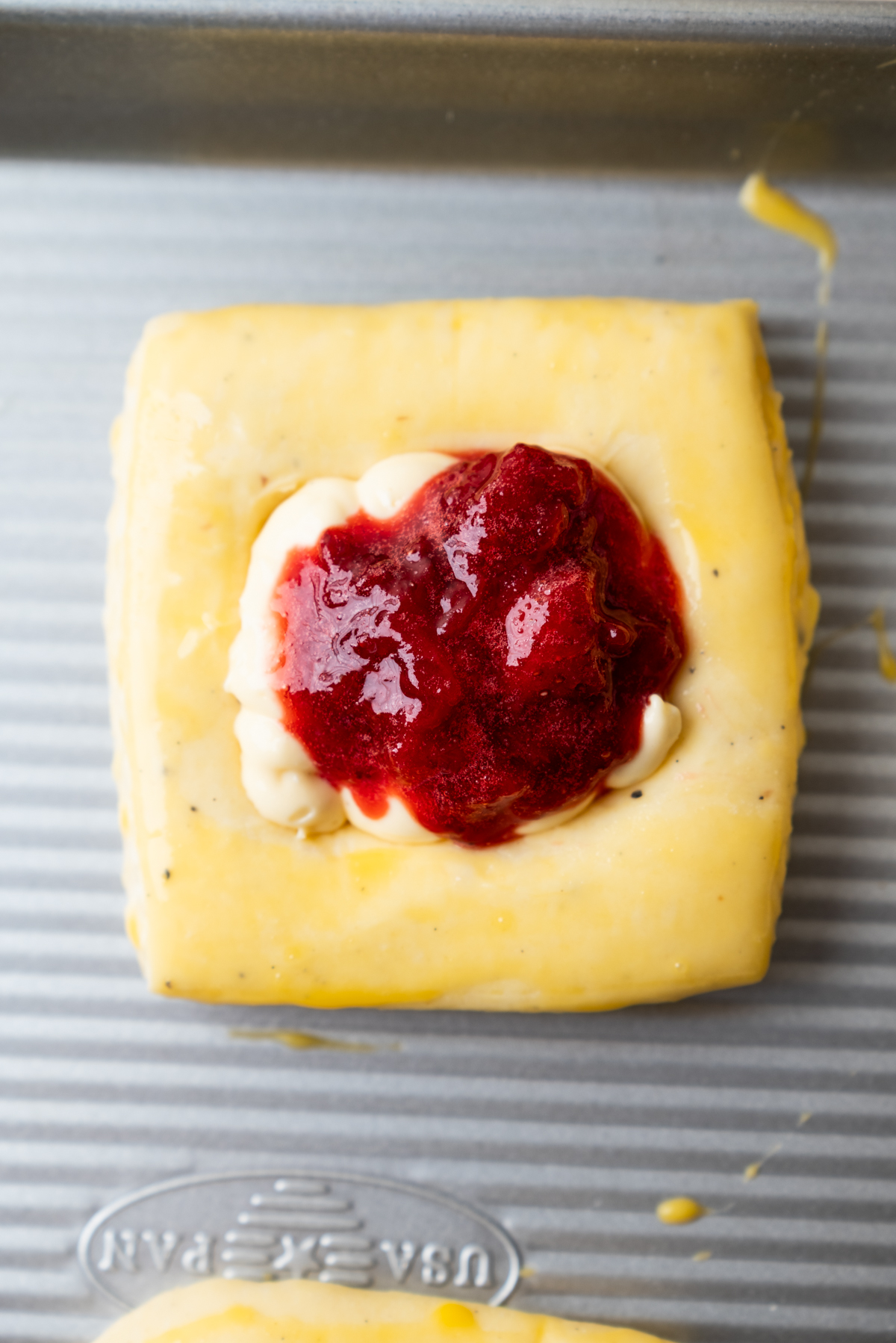 A square piece of raw pastry dough with cream cheese and a dollop of red strawberry jam in the center sits on a metal baking sheet.