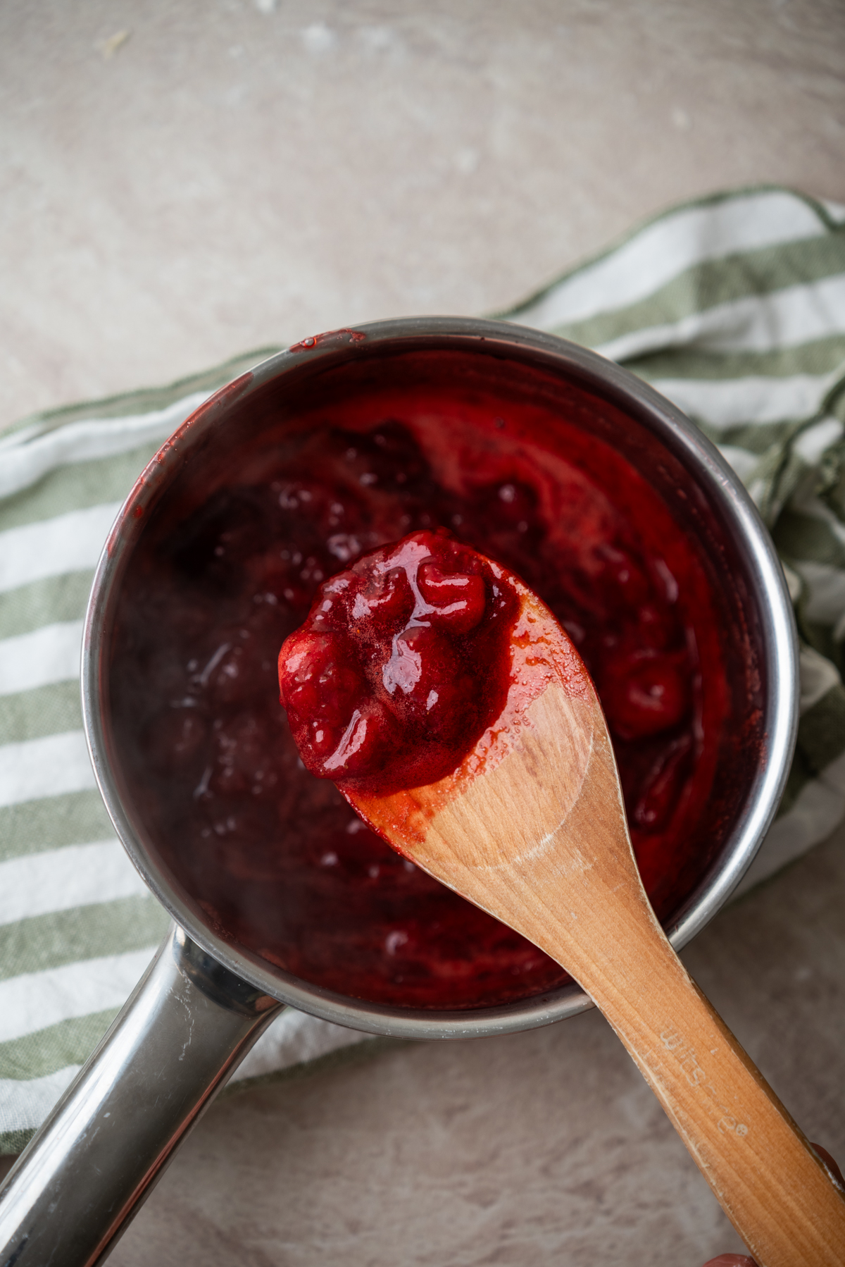 A wooden spoon holds up a thick red fruit sauce over a saucepan, with a green and white striped cloth underneath on a light countertop.