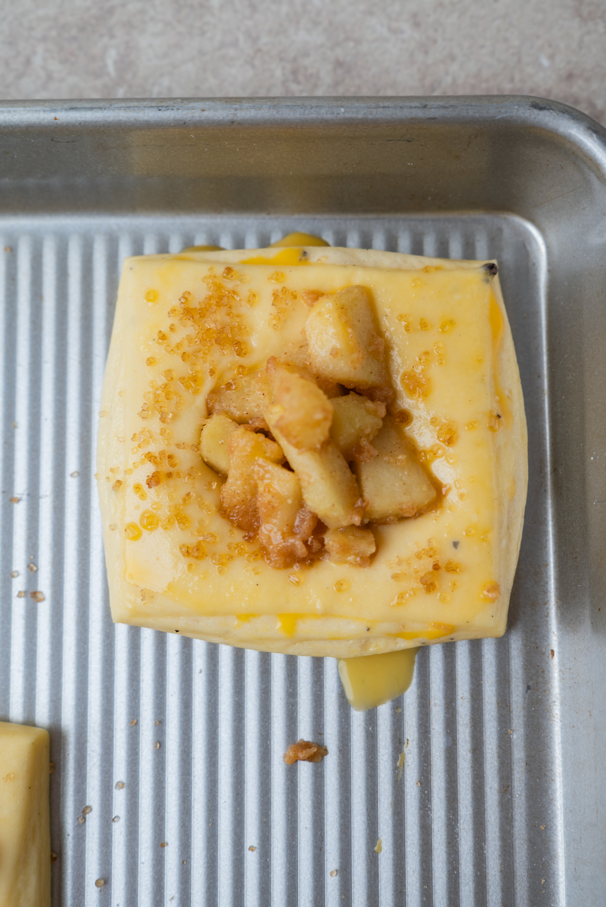 A square piece of pastry dough topped with apple chunks and brown sugar sits on a metal baking sheet, ready to be baked.