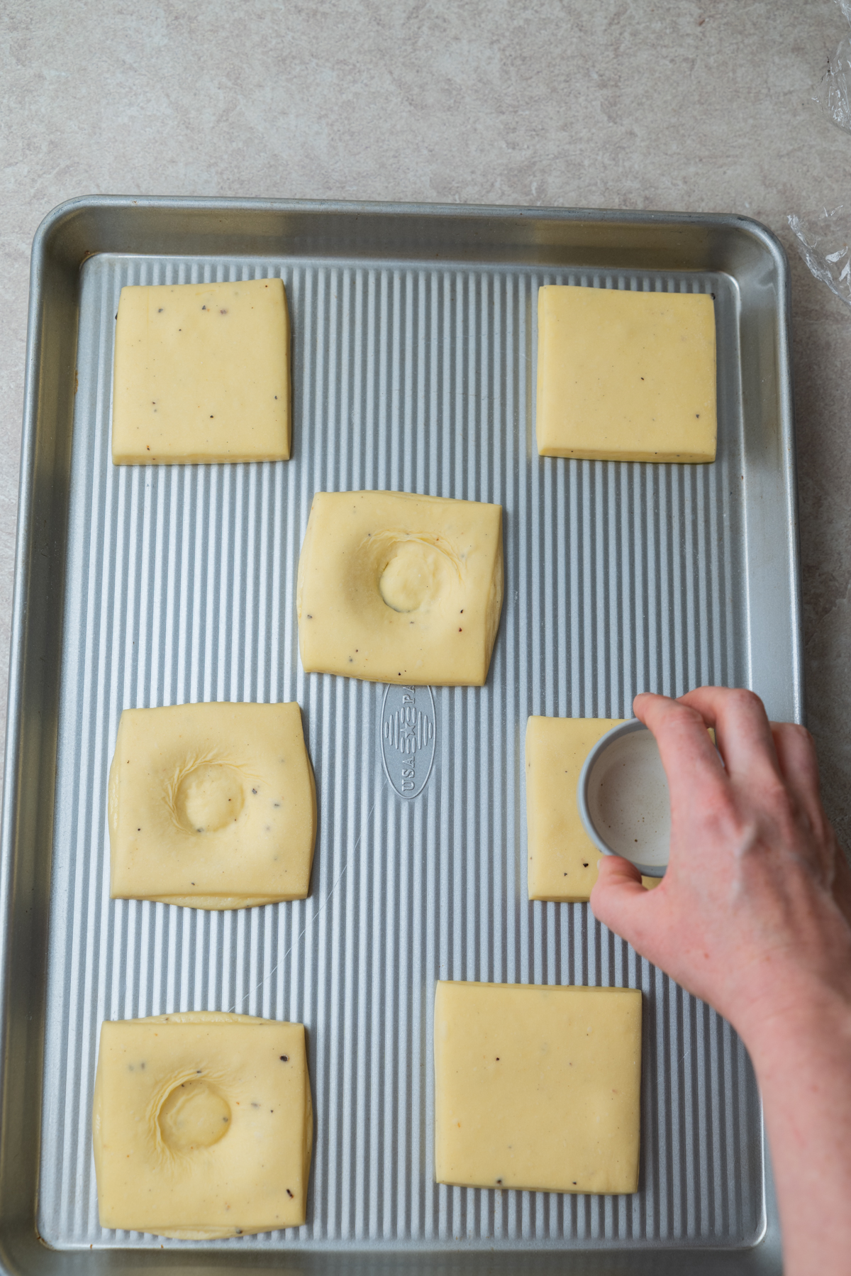 A hand presses a round object into square pieces of dough on a baking sheet, creating indentations in the center of three squares.