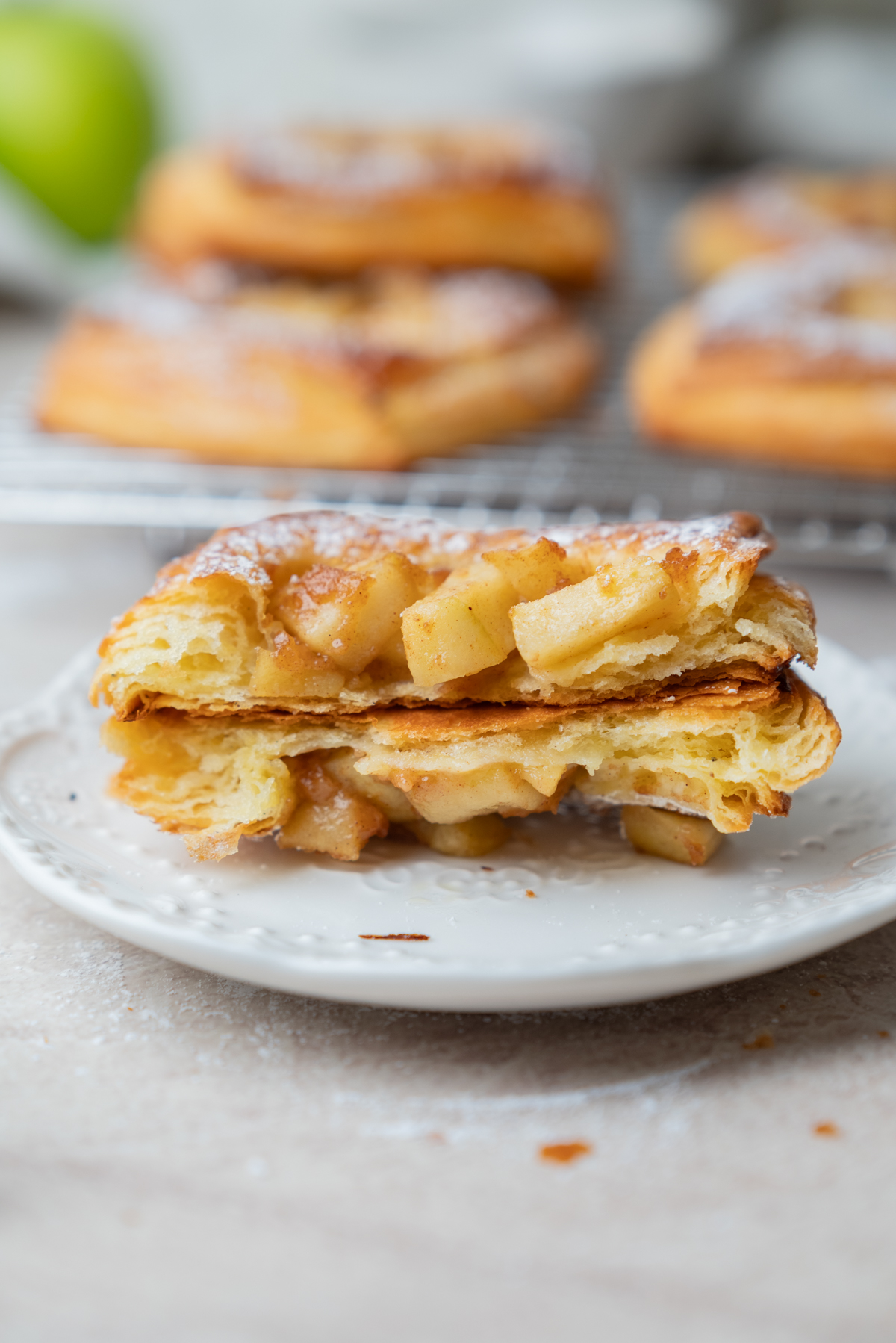 A flaky pastry filled with diced apples is cut in half and stacked on a white plate, with more pastries blurred in the background.