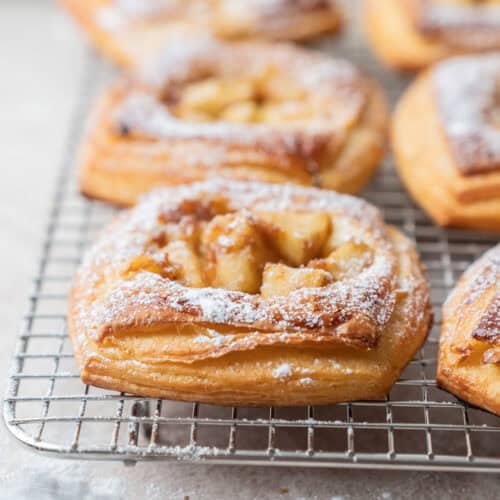 Close-up of golden, flaky Strawberry Cream Cheese Danish Pastries topped with powdered sugar and apple pieces, cooling on a wire rack. A metal sifter with a bit of sugar rests in the corner on a light countertop.
