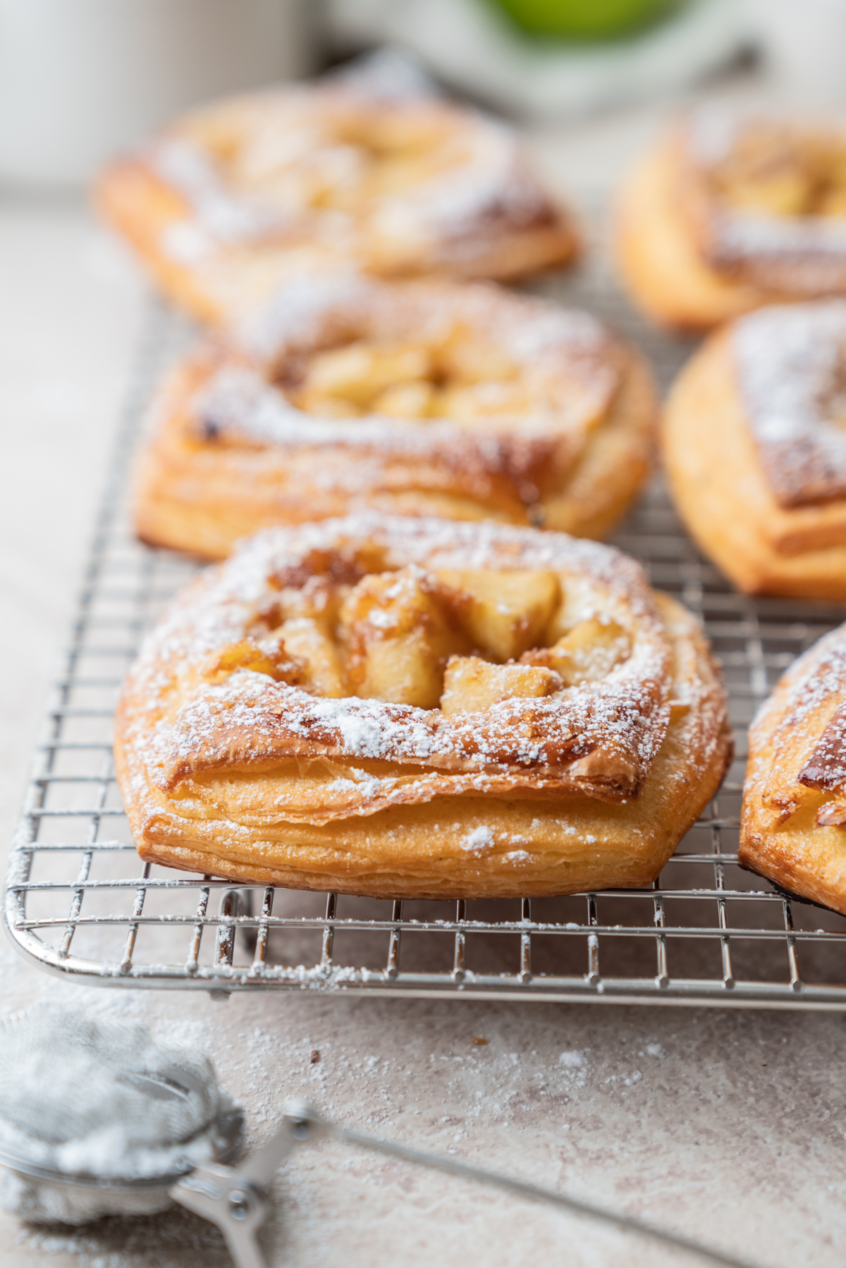 Close-up of golden, flaky Strawberry Cream Cheese Danish Pastries topped with powdered sugar and apple pieces, cooling on a wire rack. A metal sifter with a bit of sugar rests in the corner on a light countertop.