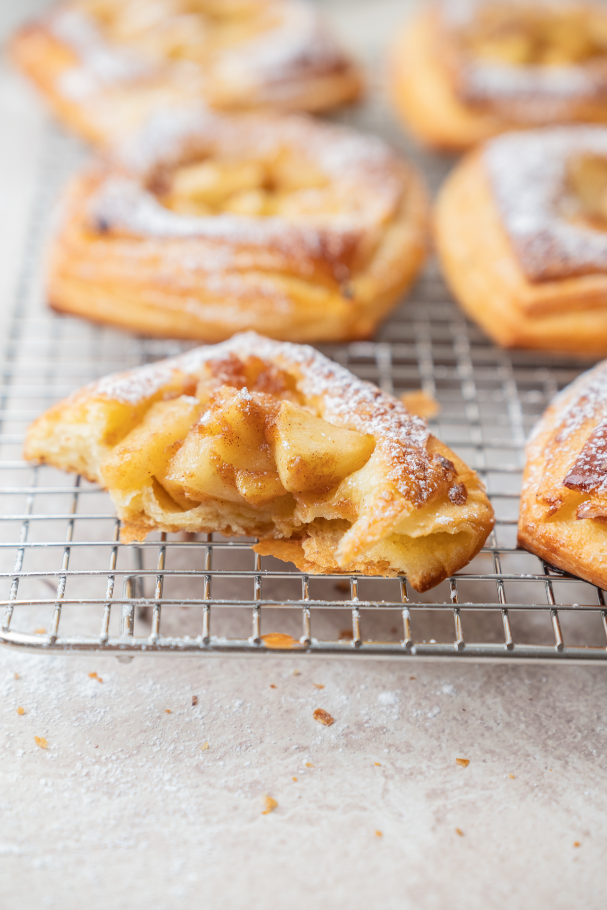 A close-up of apple danish pastries on a cooling rack, with one pastry showing a bite taken out, revealing spiced apple filling. Nestled beside them are Strawberry Cream Cheese Danish Pastries, their golden layers and creamy centers beckoning. All the pastries are dusted with powdered sugar for a sweet finishing touch.