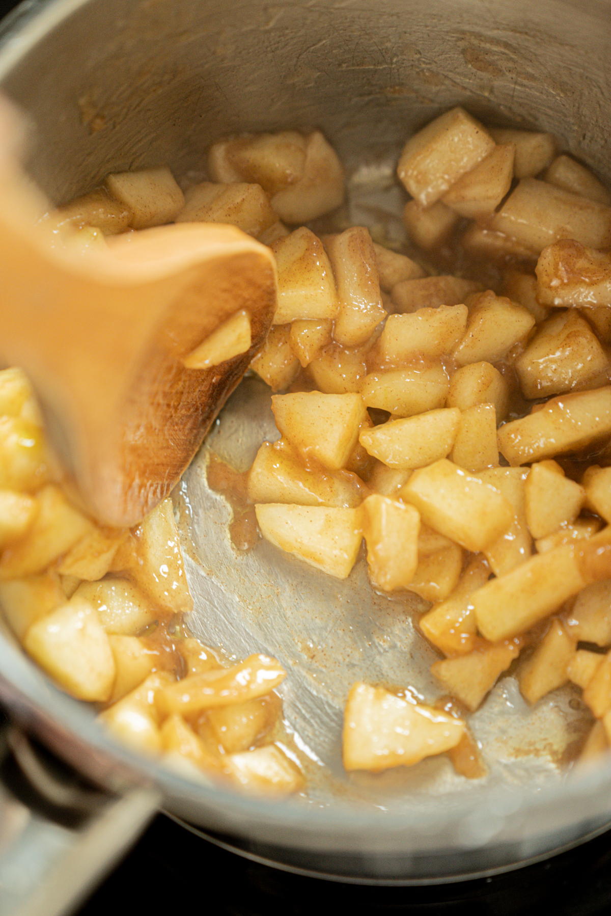 Chopped apples being stirred with a wooden spoon in a pot, coated in a glossy, cinnamon-spiced sauce—much like the luscious fillings you might find tucked inside Strawberry Cream Cheese Danish Pastries—possibly being prepared for a pie or dessert.