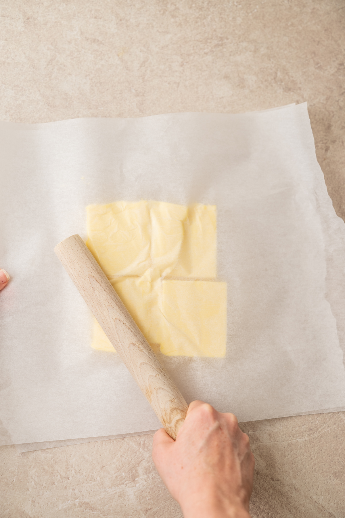 A hand uses a rolling pin to flatten a square of butter between two sheets of parchment paper on a light countertop.