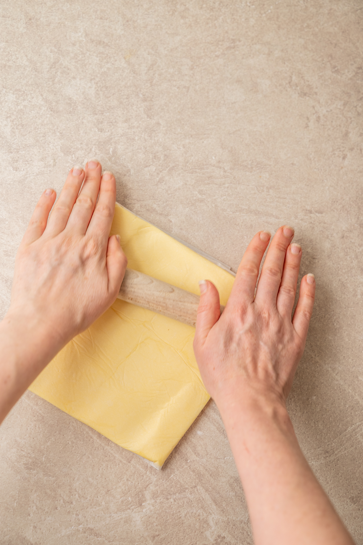 Two hands roll up a sheet of pale dough on a light, textured countertop.