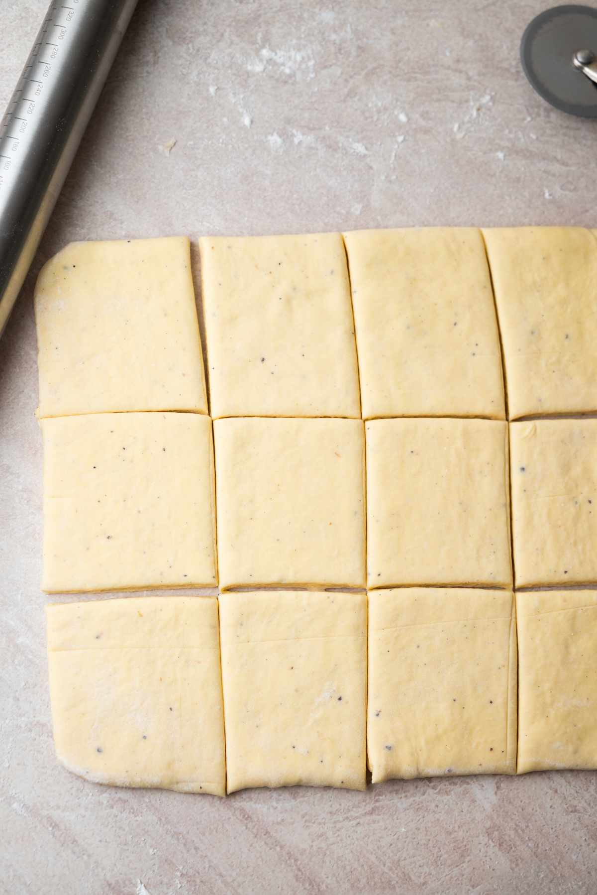 A sheet of dough cut into twelve equal squares sits on a floured surface, with a rolling pin and a pizza cutter nearby.