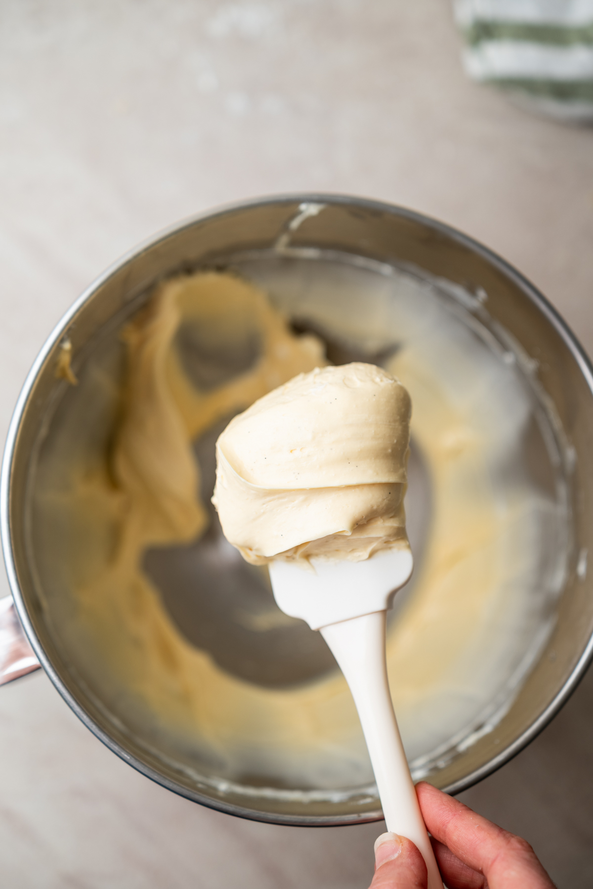 A hand holds a white spatula with creamy frosting above a metal mixing bowl containing more of the same frosting.