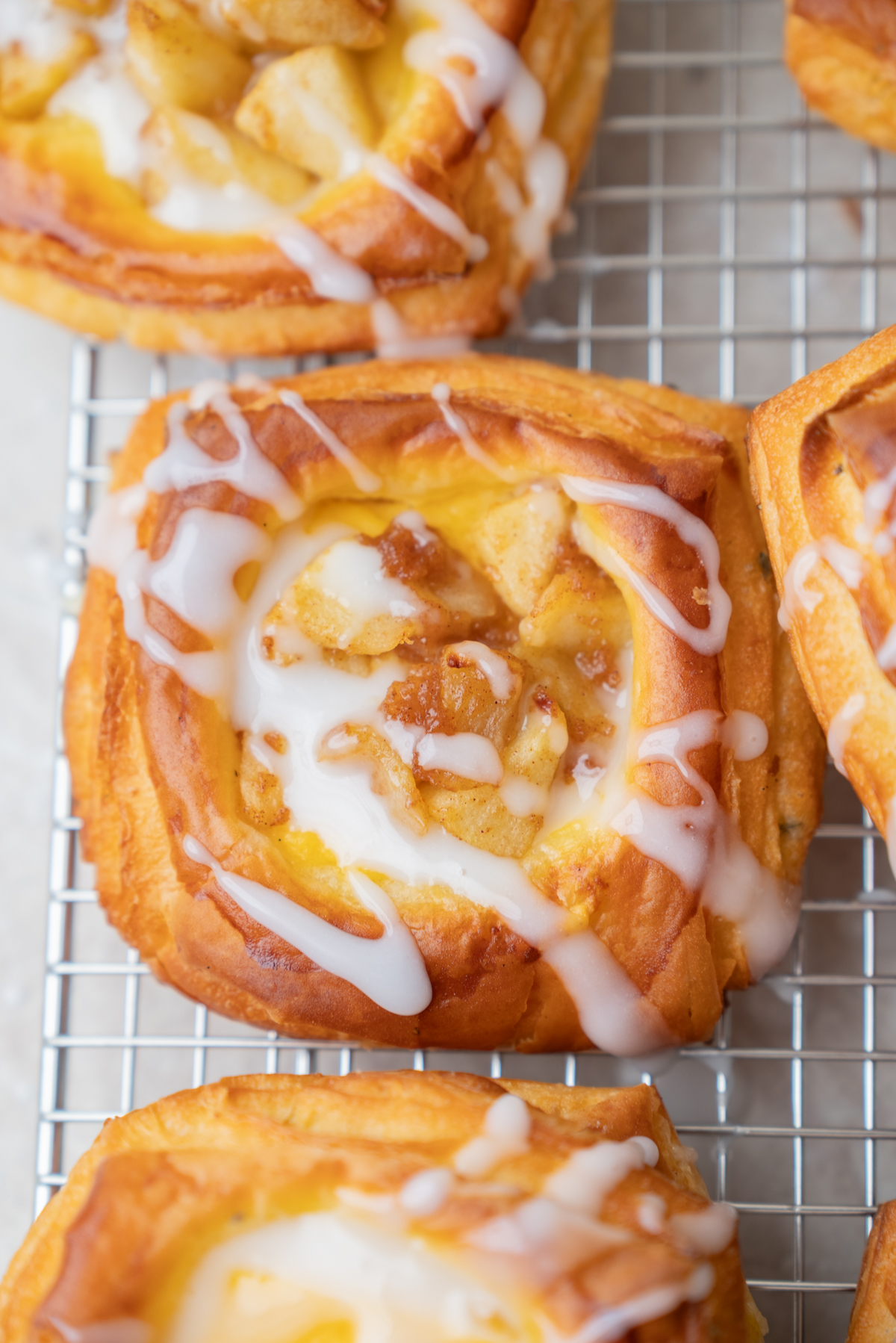 A close-up of a square Danish pastry topped with icing and small apple pieces, resting on a wire cooling rack.
