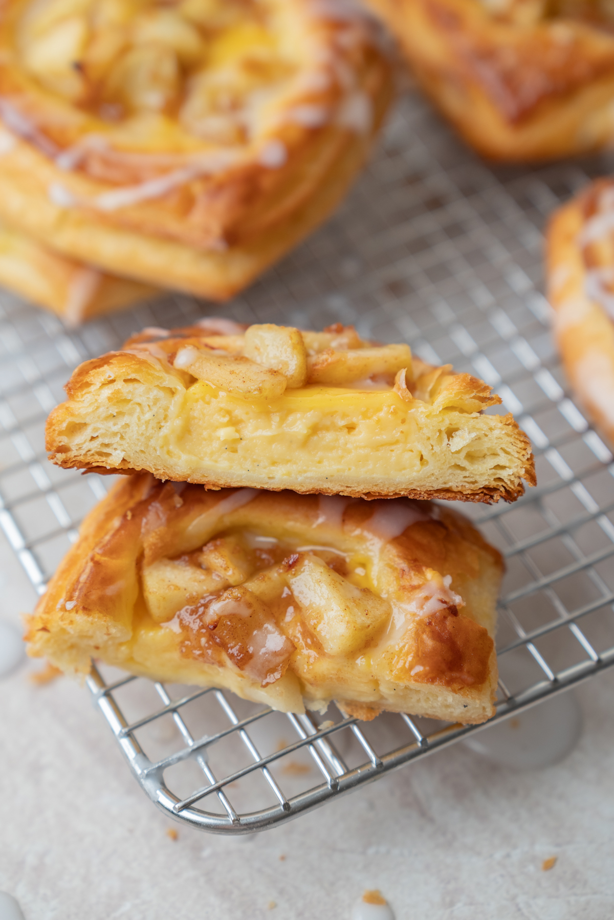 Two pieces of apple danish with icing and custard are stacked on a cooling rack. The pastry is flaky and golden, with pieces of baked apple and a drizzle of icing visible on top.