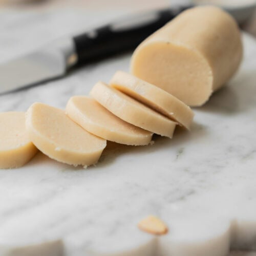 A log of marzipan is partially sliced on a marble board, with a knife and a small dish in the background.