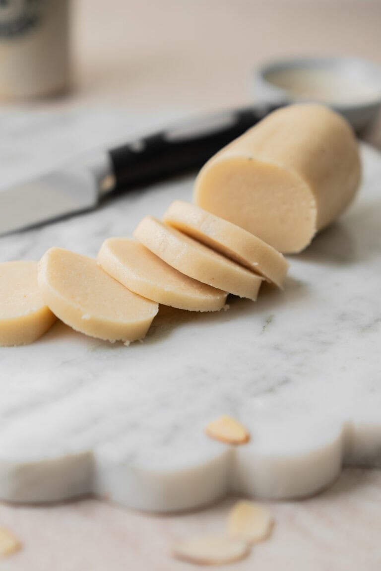 A log of marzipan is partially sliced on a marble board, with a knife and a small dish in the background.