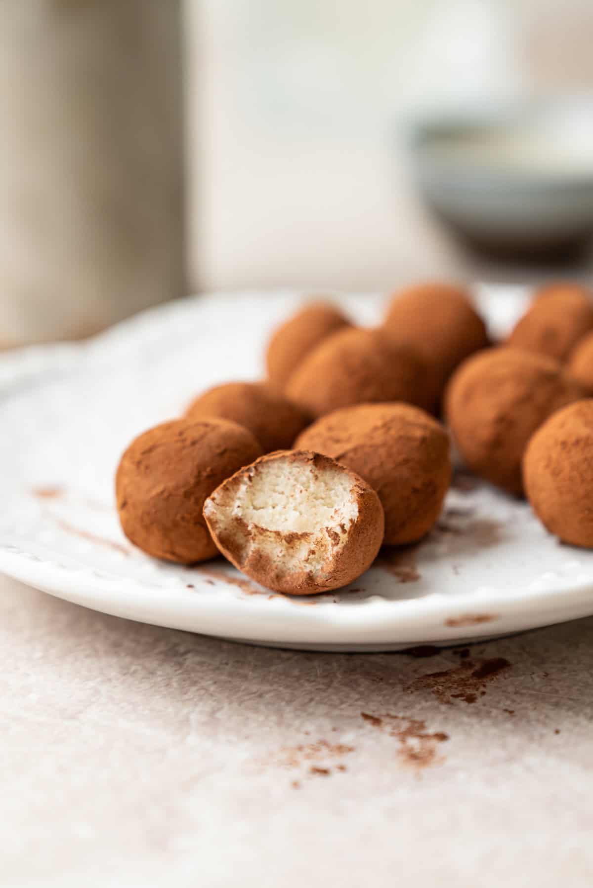 A white plate holds several cocoa-dusted truffles, one of which has a bite taken out, revealing a creamy filling inside. The background is softly blurred.
