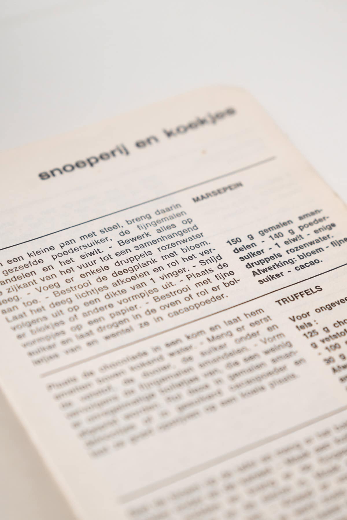 A close-up of a Dutch recipe book showing a page with text and headings for MANDELEN and TRUFFELS. The focus is on the printed words, with the page angled against a white background.
