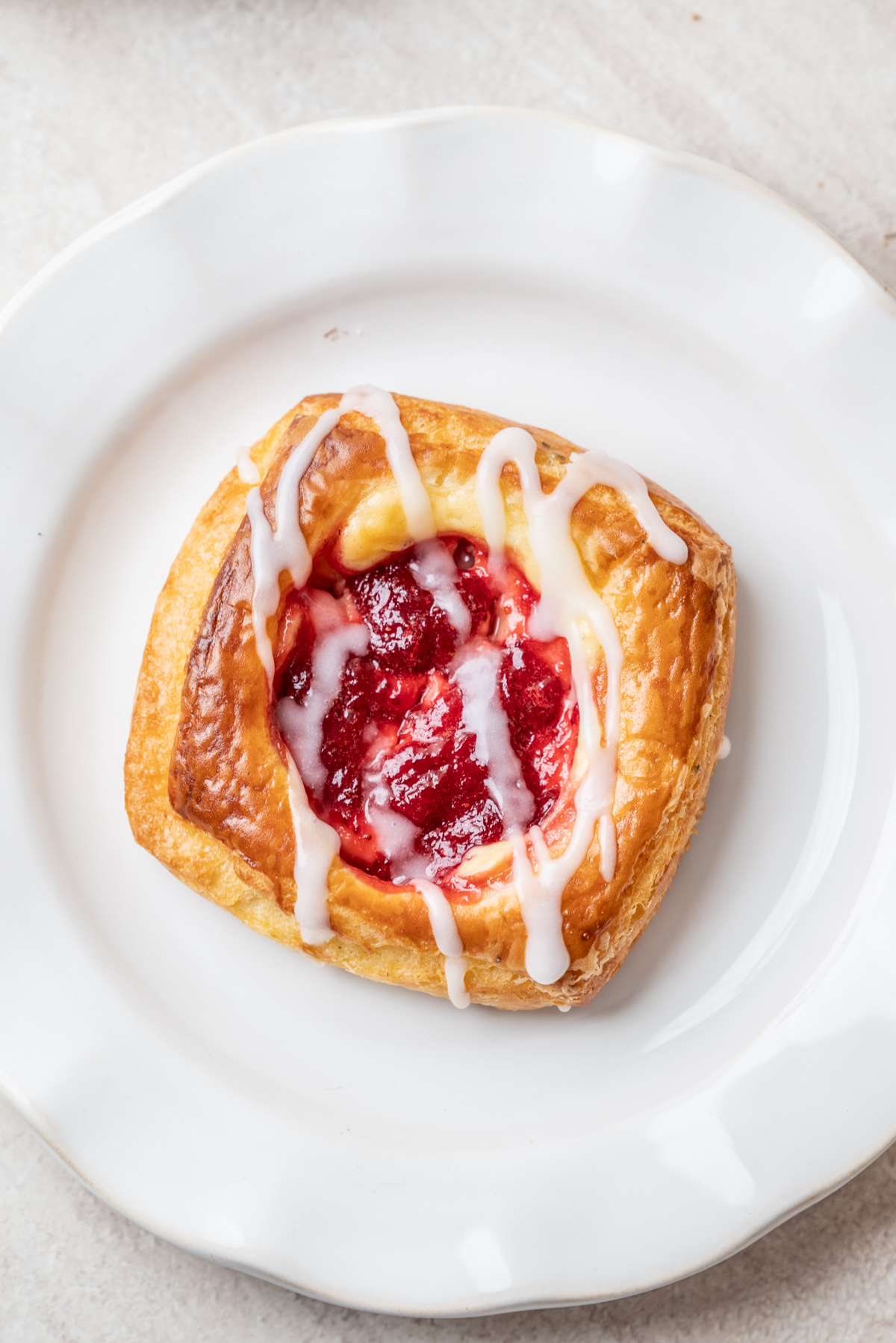A square pastry with golden crust, filled with red fruit jam and drizzled with white icing, sits on a white plate.