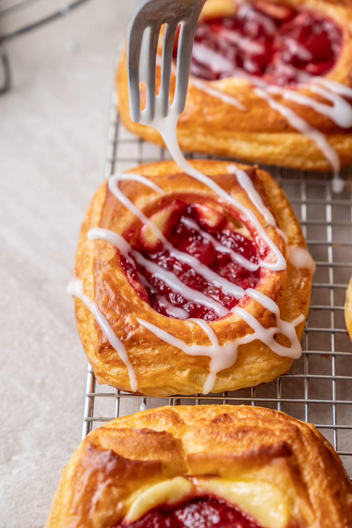 A fork hovers over a fruit Danish pastry drizzled with white icing, resting on a cooling rack.