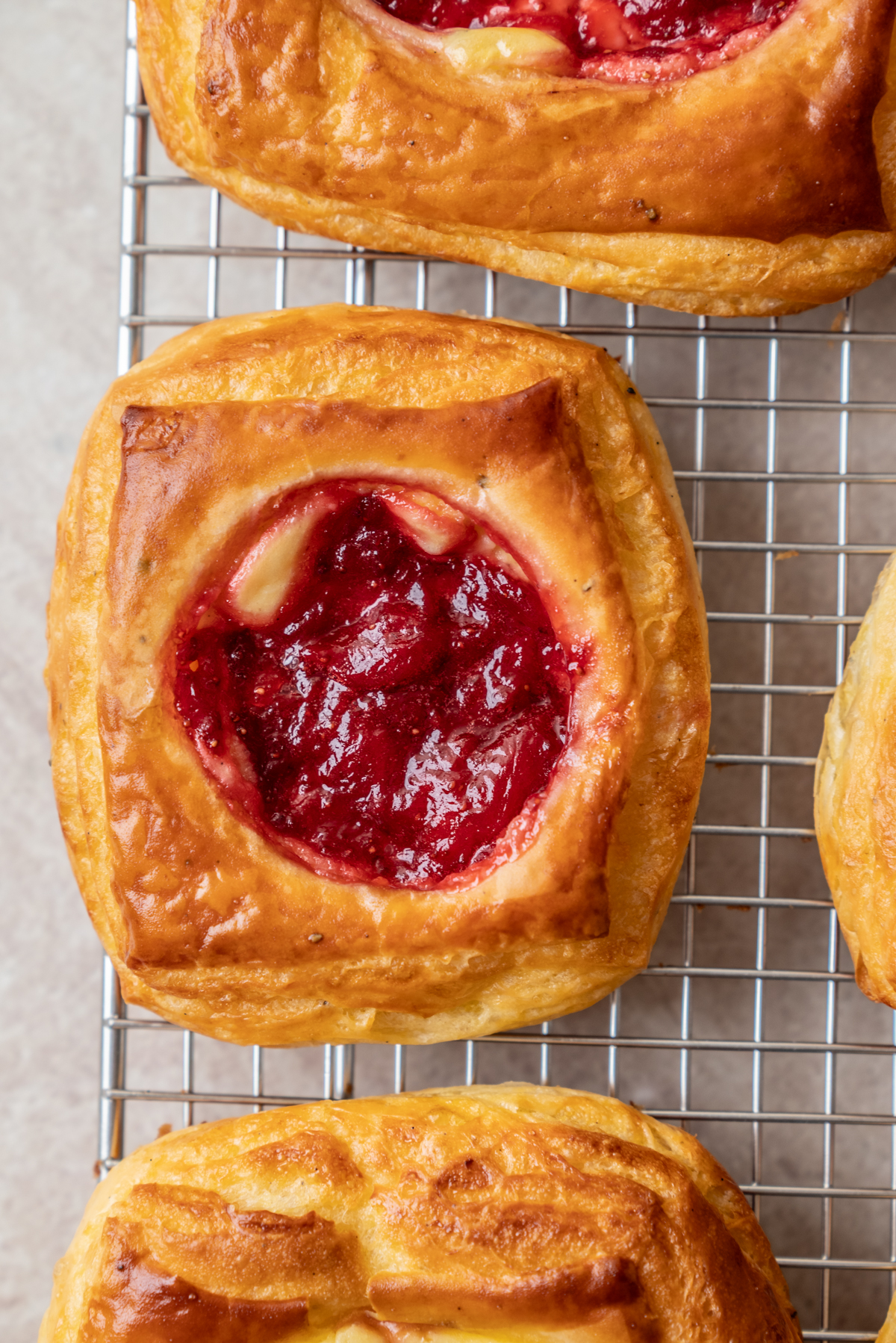 A close-up of square pastries with golden, flaky crusts and a center filled with bright red fruit jam, cooling on a metal wire rack.