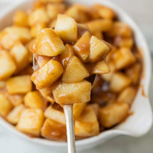 A spoonful of cooked, cinnamon-coated brown butter apple pie filling apple chunks held over a white bowl filled with more of the same apple mixture.