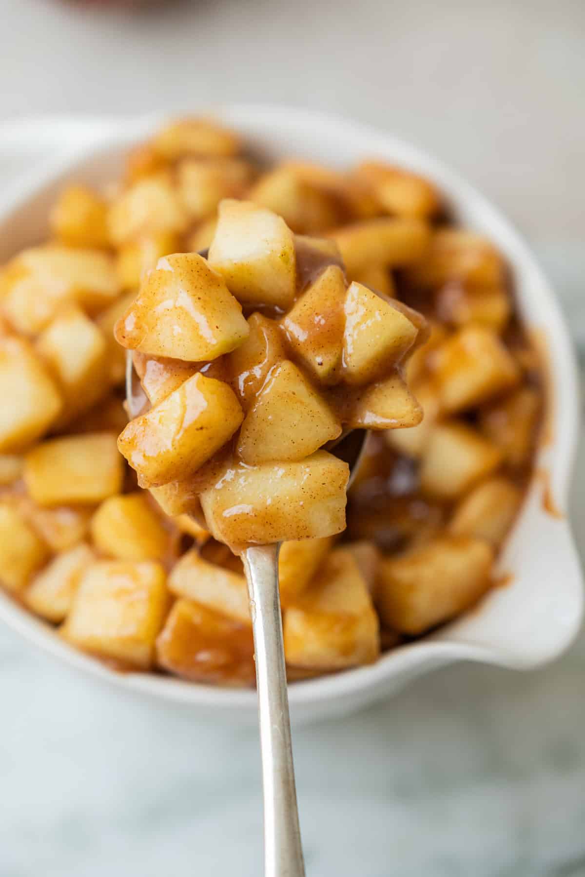 A spoonful of cooked, cinnamon-coated brown butter apple pie filling apple chunks held over a white bowl filled with more of the same apple mixture.