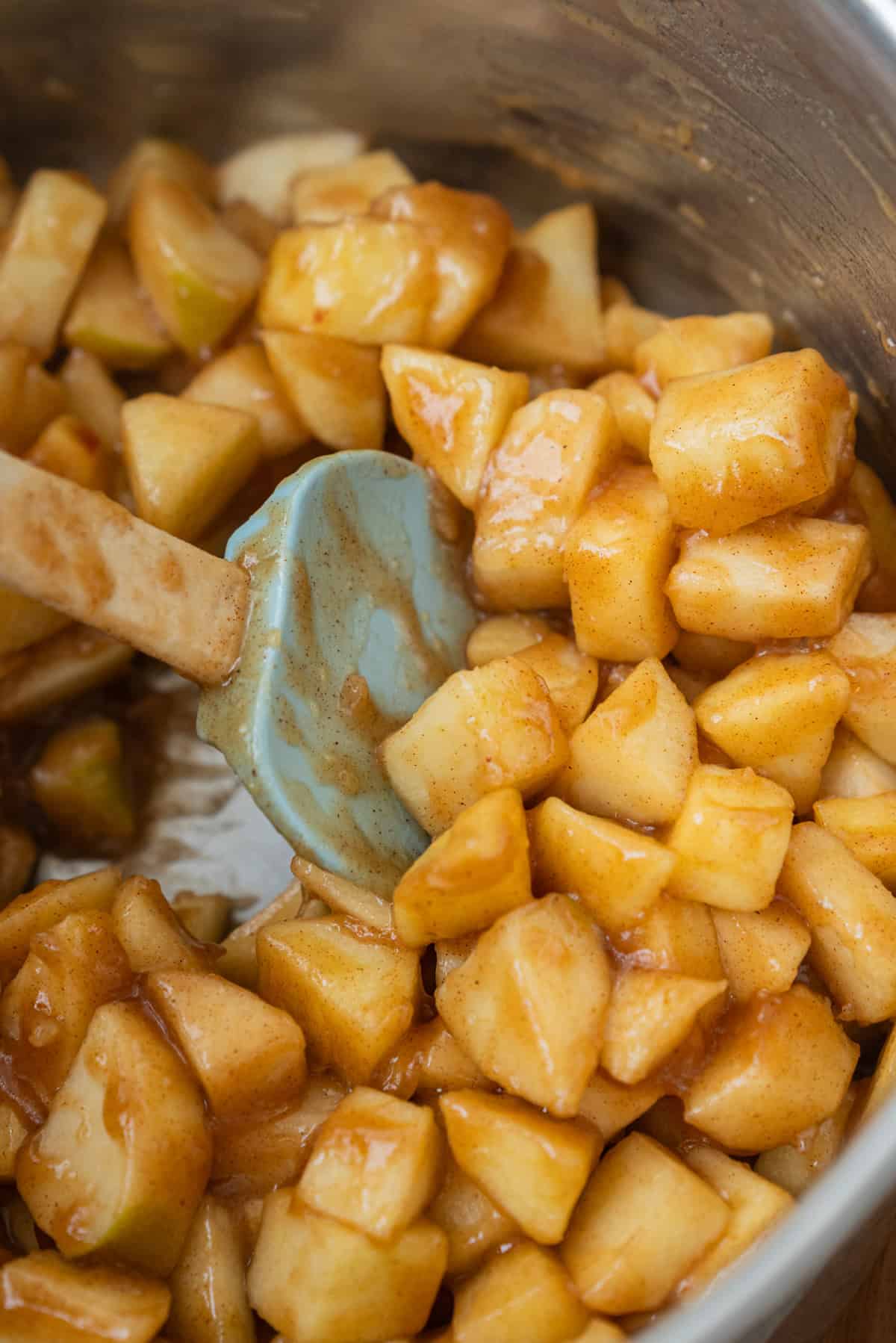 A blue silicone spatula stirring diced apples coated in a cinnamon glaze in a metal pot.