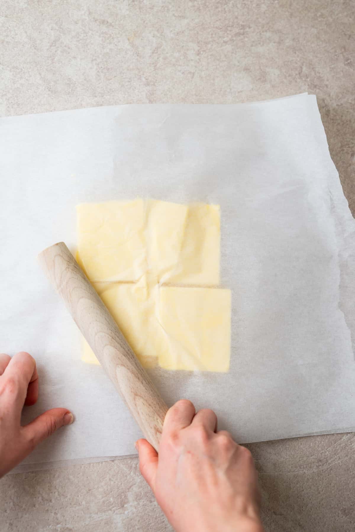 Hands use a wooden rolling pin to flatten squares of butter between two sheets of parchment paper.