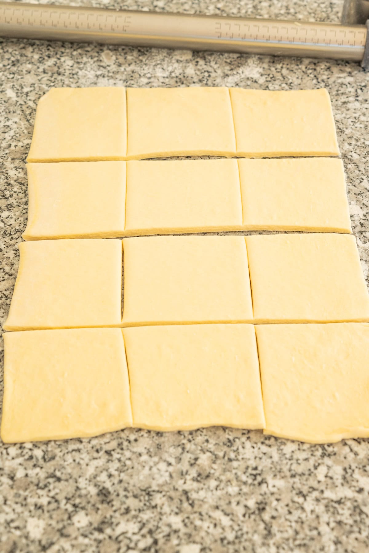 Twelve squares of rolled dough arranged in a grid on a granite countertop.