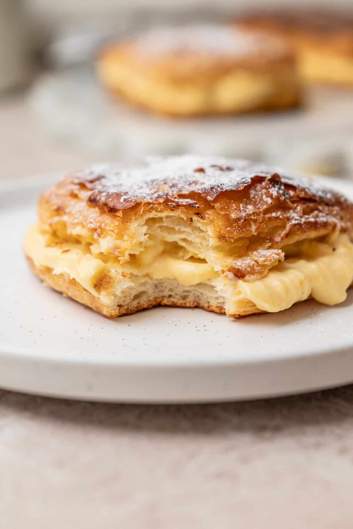 A flaky pastry filled with cream and dusted with powdered sugar, with a bite taken out of the center, resting on a white plate.