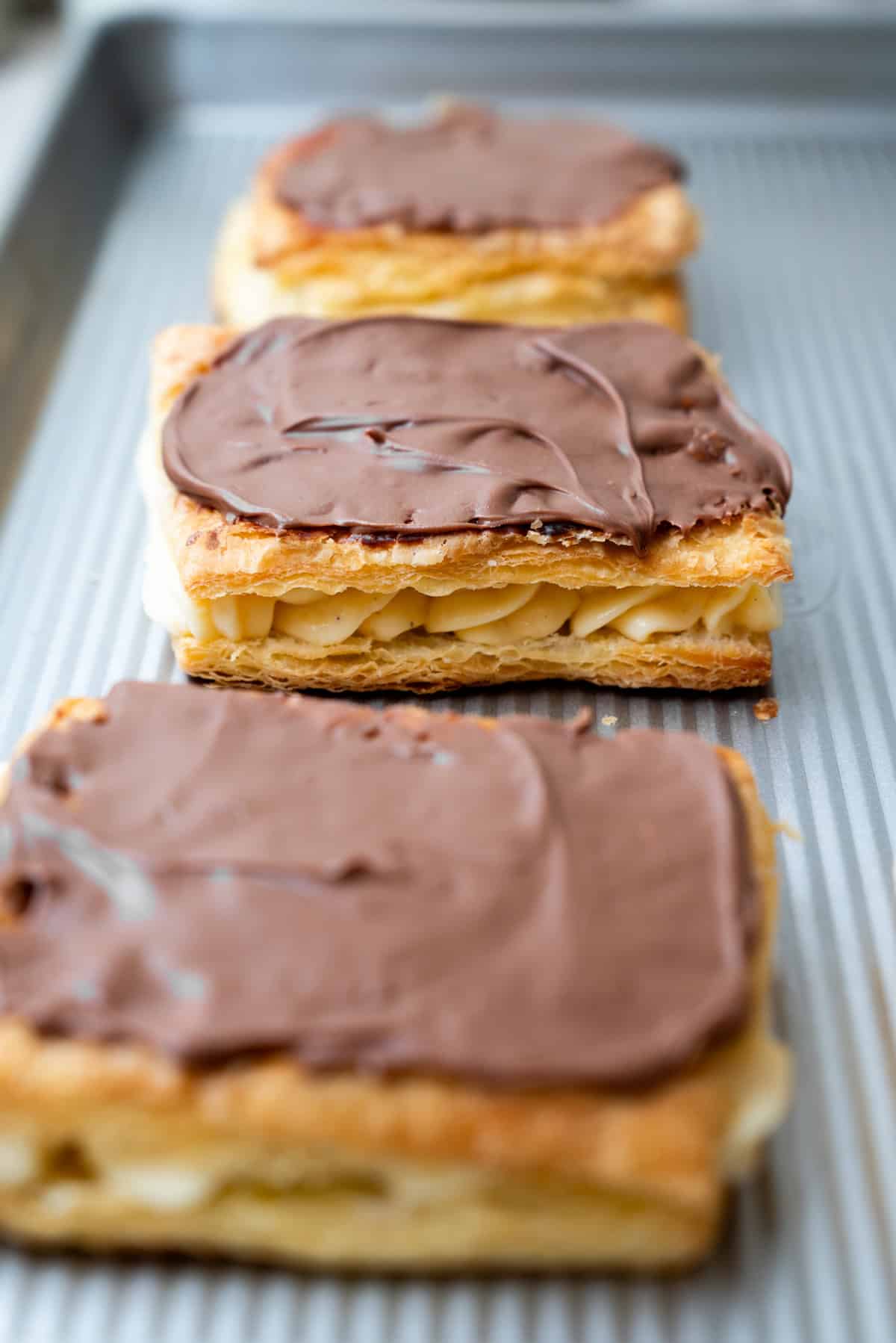 Three rectangular puff pastry desserts with vanilla cream filling and a chocolate-frosted top on a metal baking sheet.