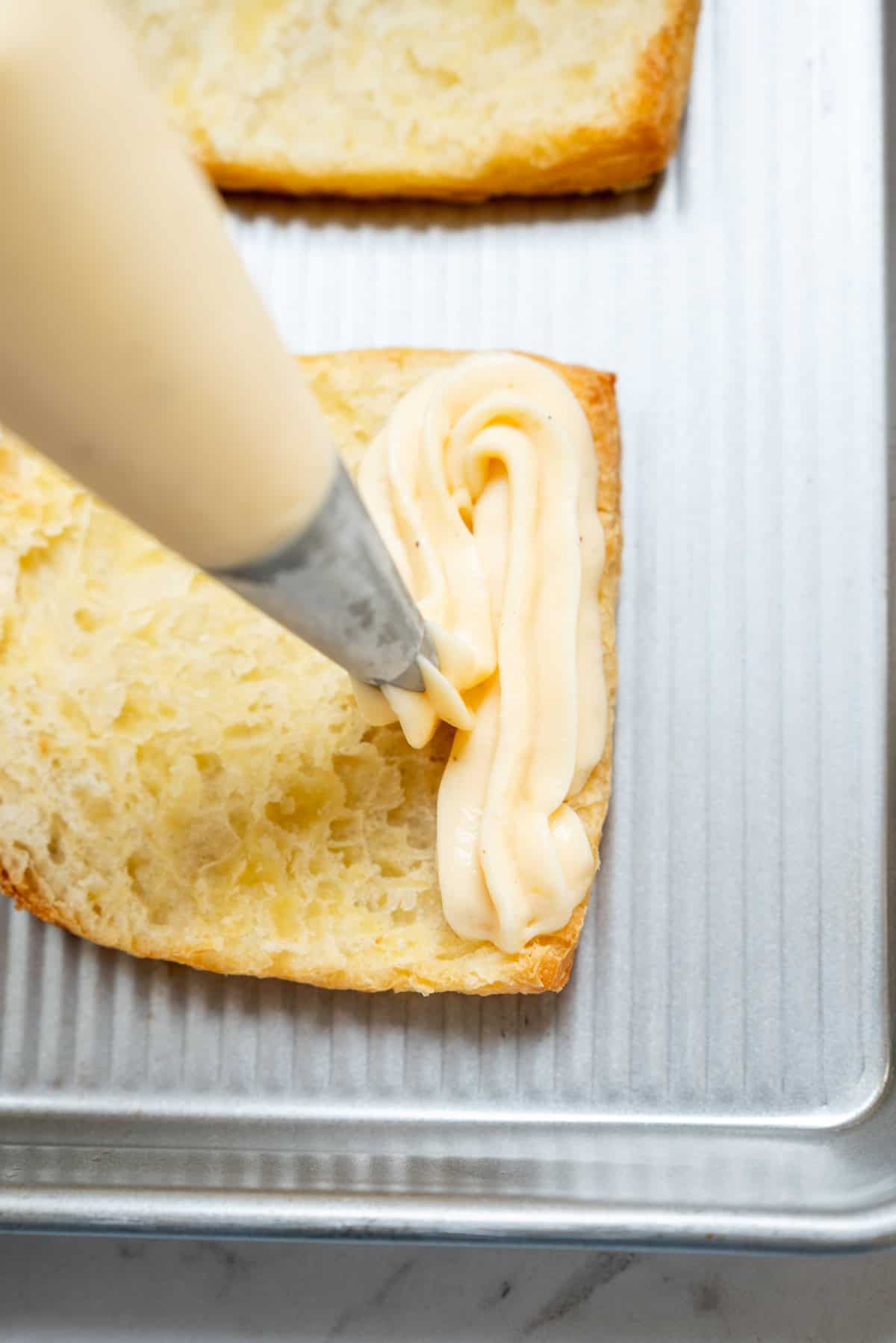 A piping bag applying cream onto a slice of bread on a baking sheet.