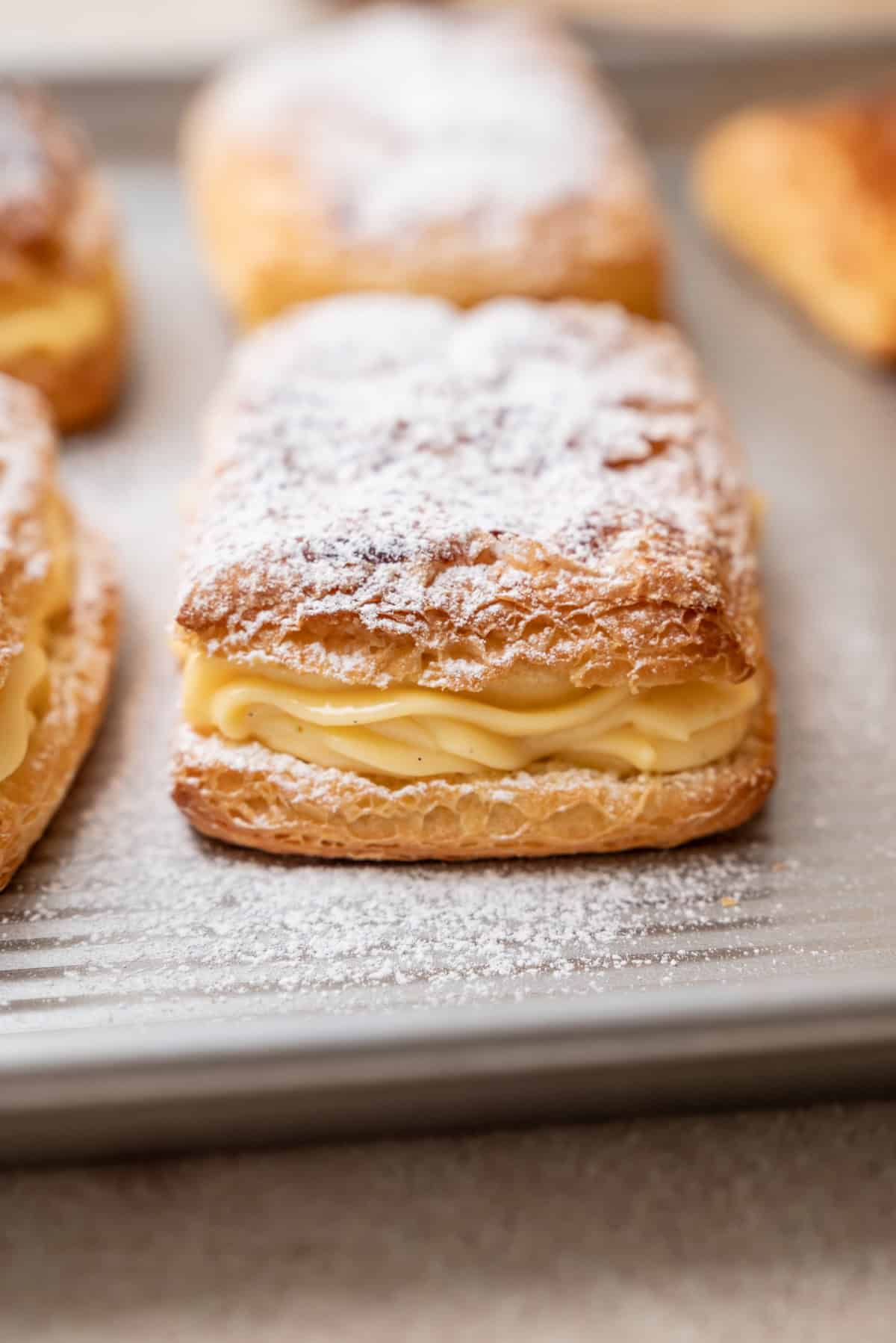 Rectangular puff pastries filled with cream and dusted with powdered sugar on a metal baking sheet.
