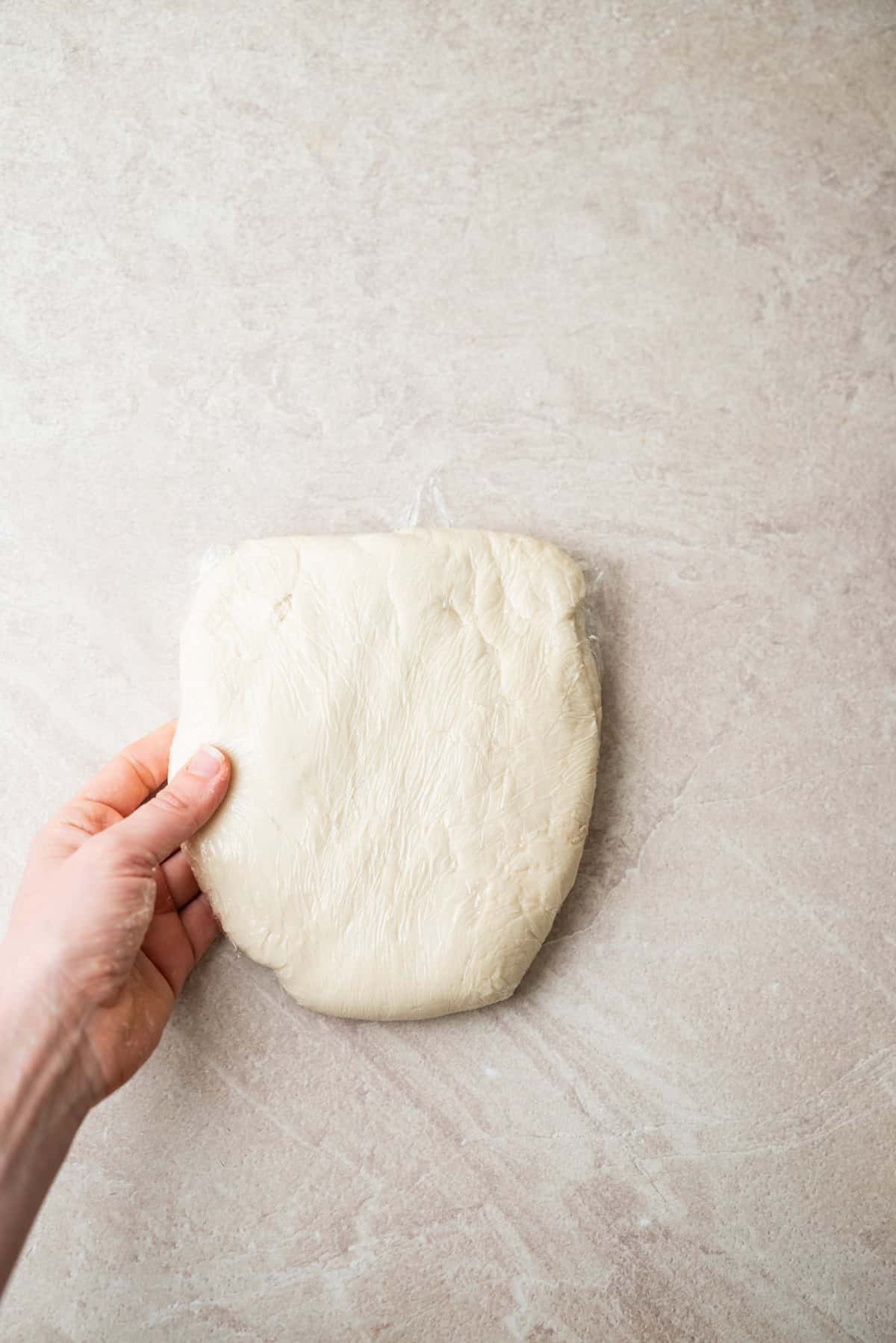 A hand holds a disk of dough wrapped in plastic wrap.