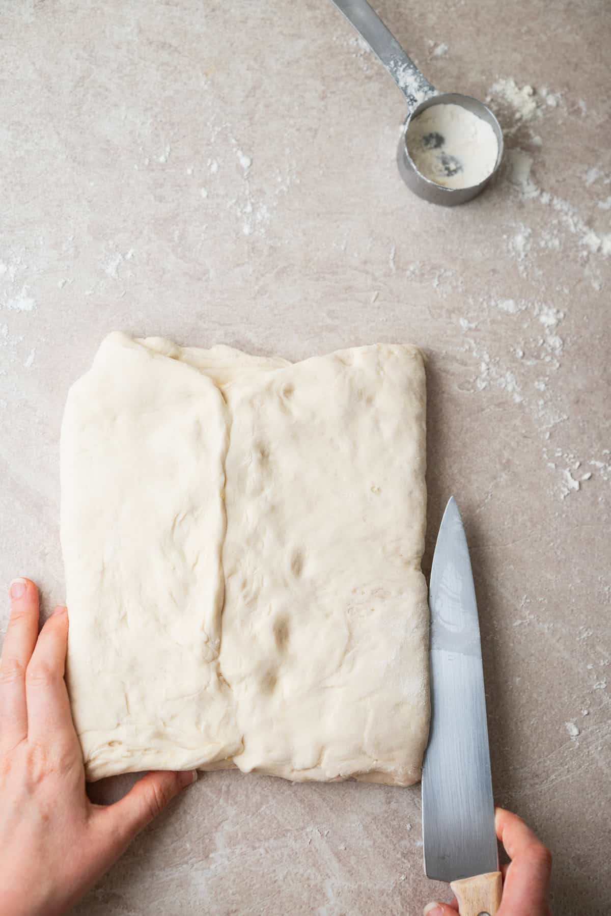Hands folding a rectangle of raw dough on a floured surface with a knife and a measuring cup nearby.