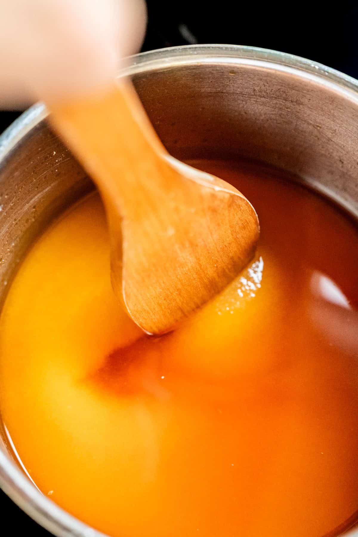 A hand stirs amber-colored liquid in a metal pot with a wooden spatula.