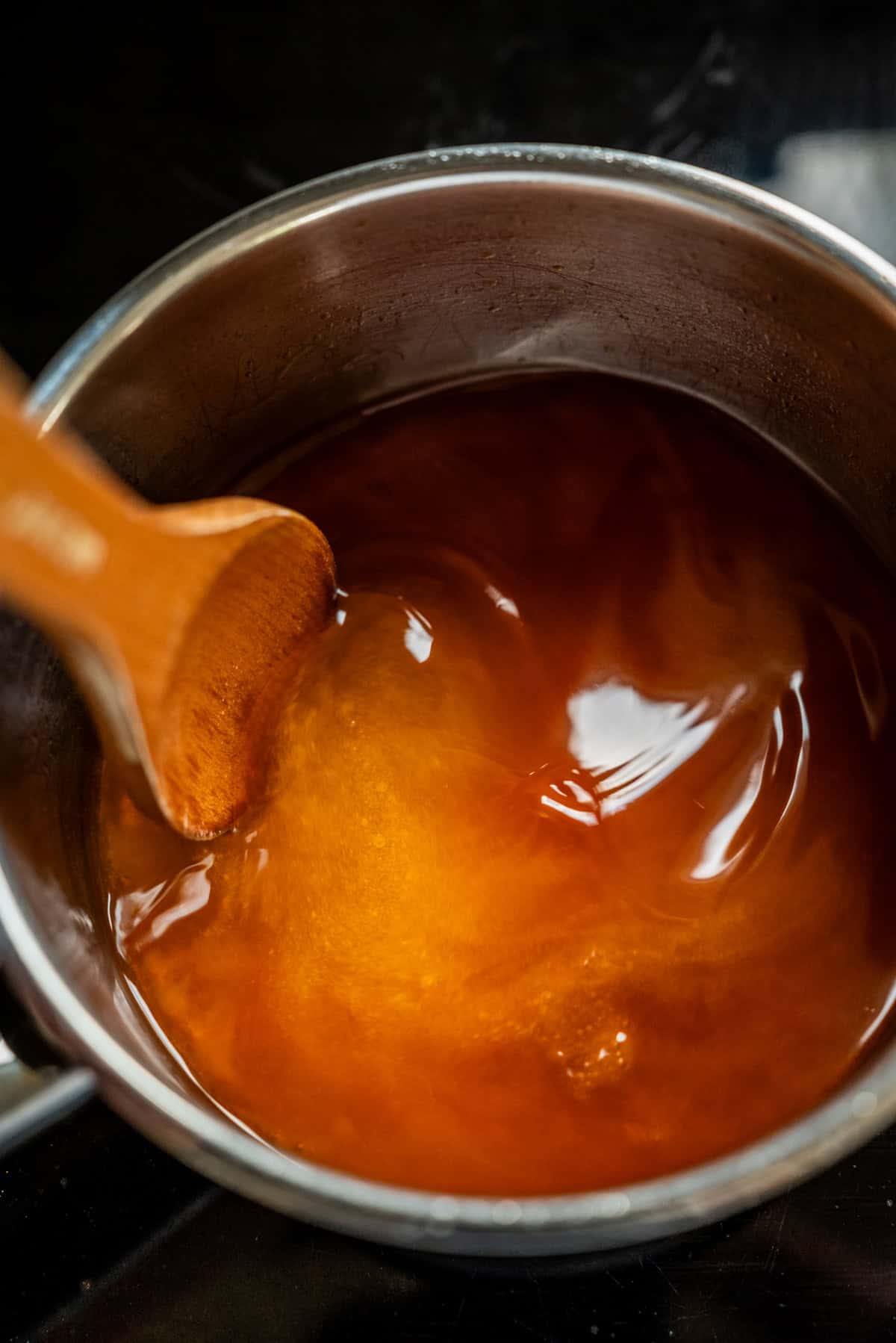 A wooden spoon stirring amber-colored liquid in a metal saucepan.